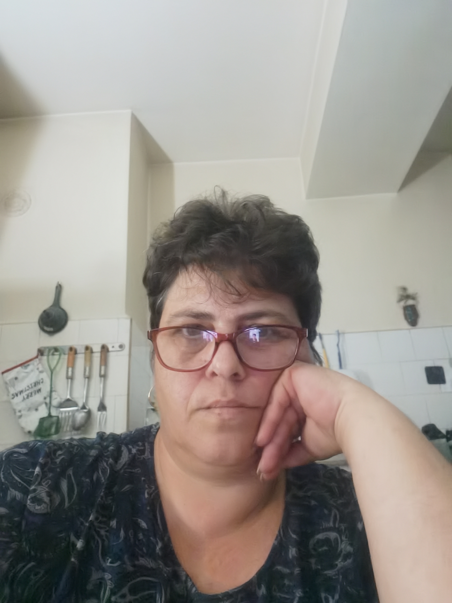 The image is a close-up selfie of a woman with short, dark, curly hair and glasses. She is wearing a dark blue shirt with a black and grey pattern. Her left hand is resting on her cheek, and her head is tilted slightly. She is in what appears to be a kitchen, with cabinets and kitchen utensils visible in the background. The lighting suggests an indoor setting. No specific location in Lomets, Bulgaria, can be identified from the image.
