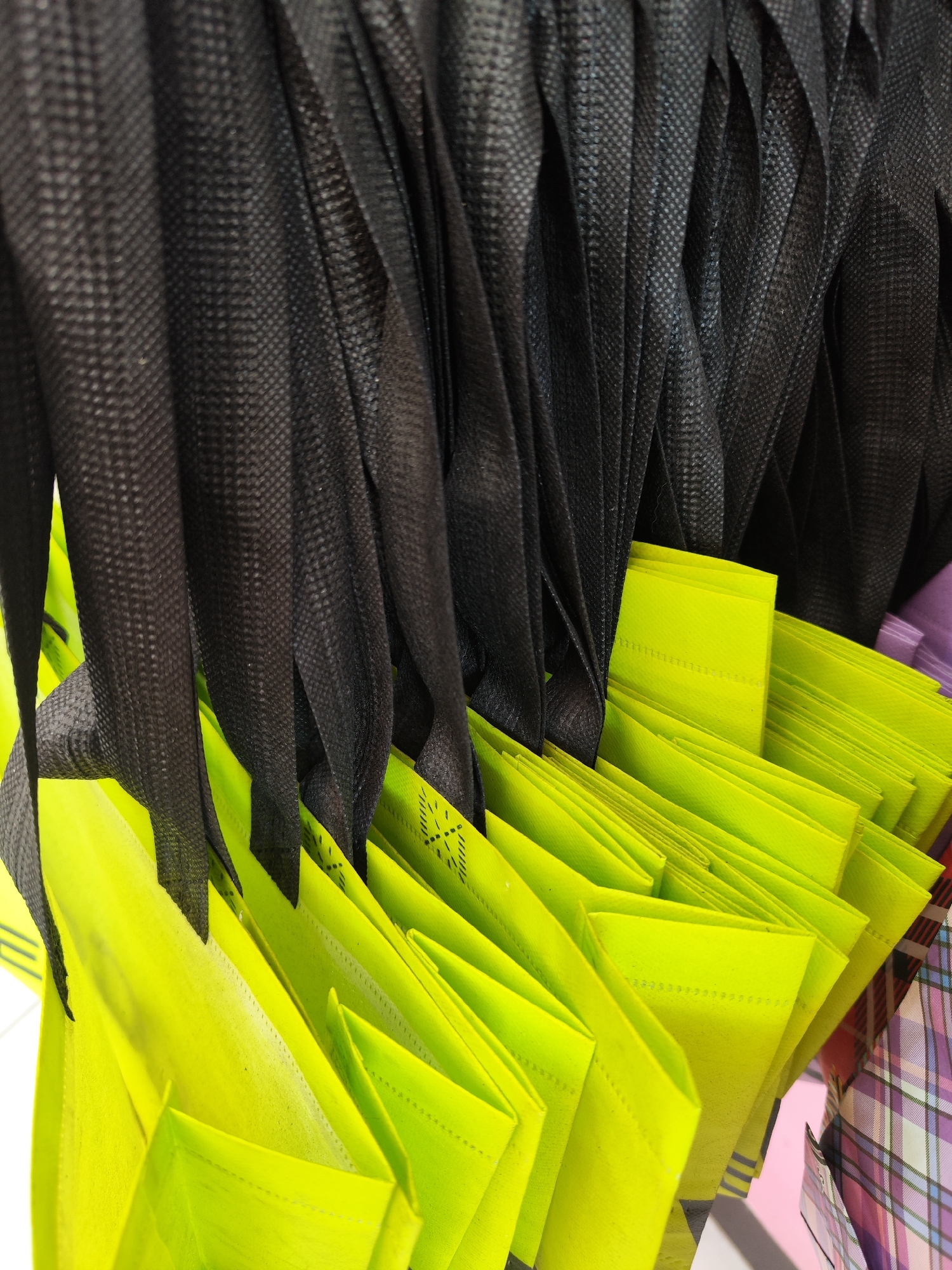 The image shows a close-up, abstract view of folded non-woven bags. The bags are primarily bright lime green and black. The green bags are stacked and folded, revealing their creased texture and stitch lines. Some have a faint diamond pattern on them. The black material is draped above and behind the green bags, creating a contrasting backdrop of deep folds. The lighting appears to be indoor, possibly in a store or storage area, as suggested by the even illumination and the presence of other colorful folded items in the background, including what looks like plaid fabric. The image does not depict any specific recognizable landmarks or street views of Sragen, Indonesia.
