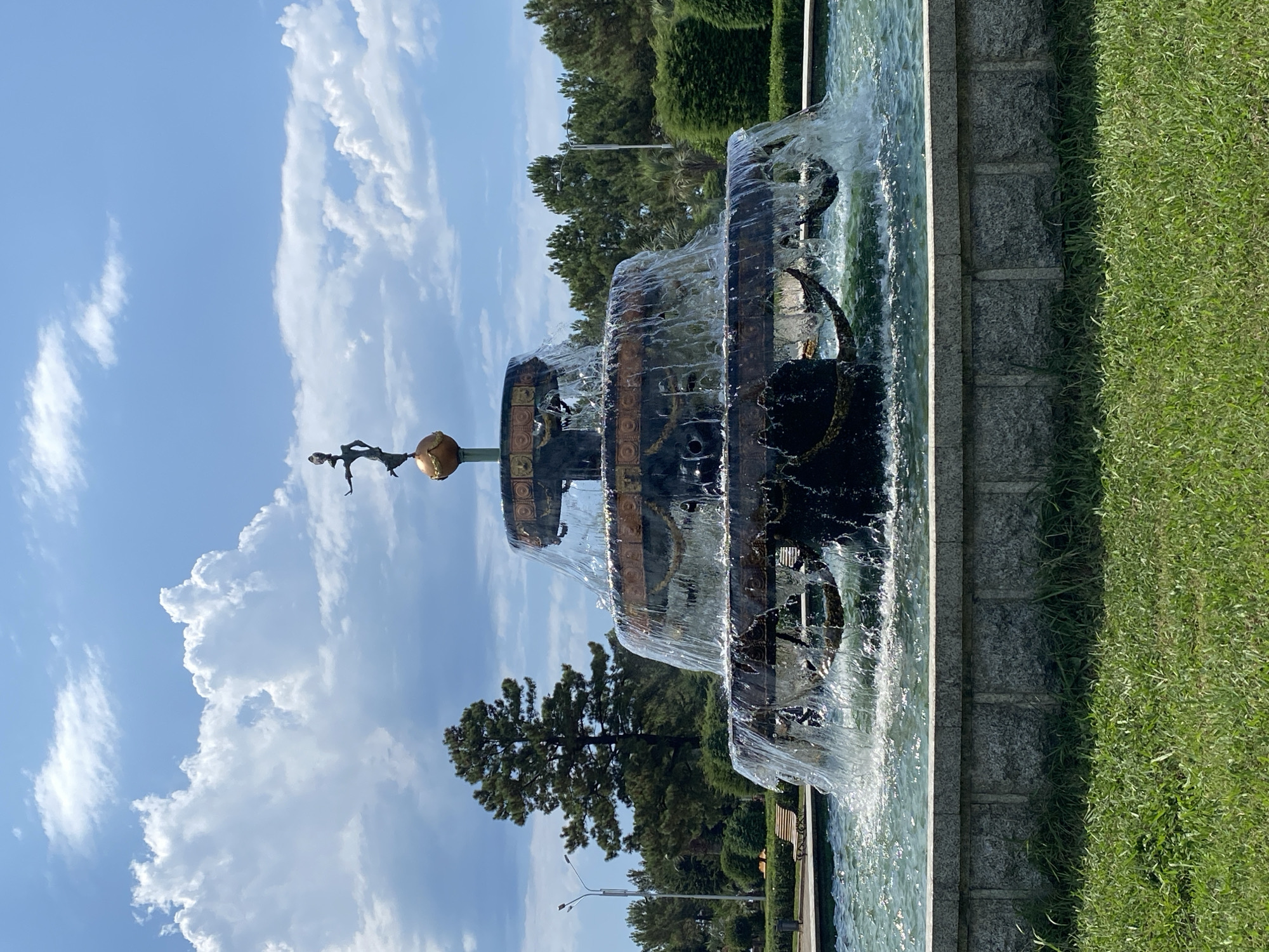 The image shows a multi-tiered fountain with water cascading down. At the very top of the fountain, there is a bronze statue of a woman standing on a globe. The fountain is surrounded by a pool of water, and the base of the fountain is made of grey stone. In the background, there are trees, lush green grass, and a blue sky with white clouds. The fountain is located in Batumi, Georgia.