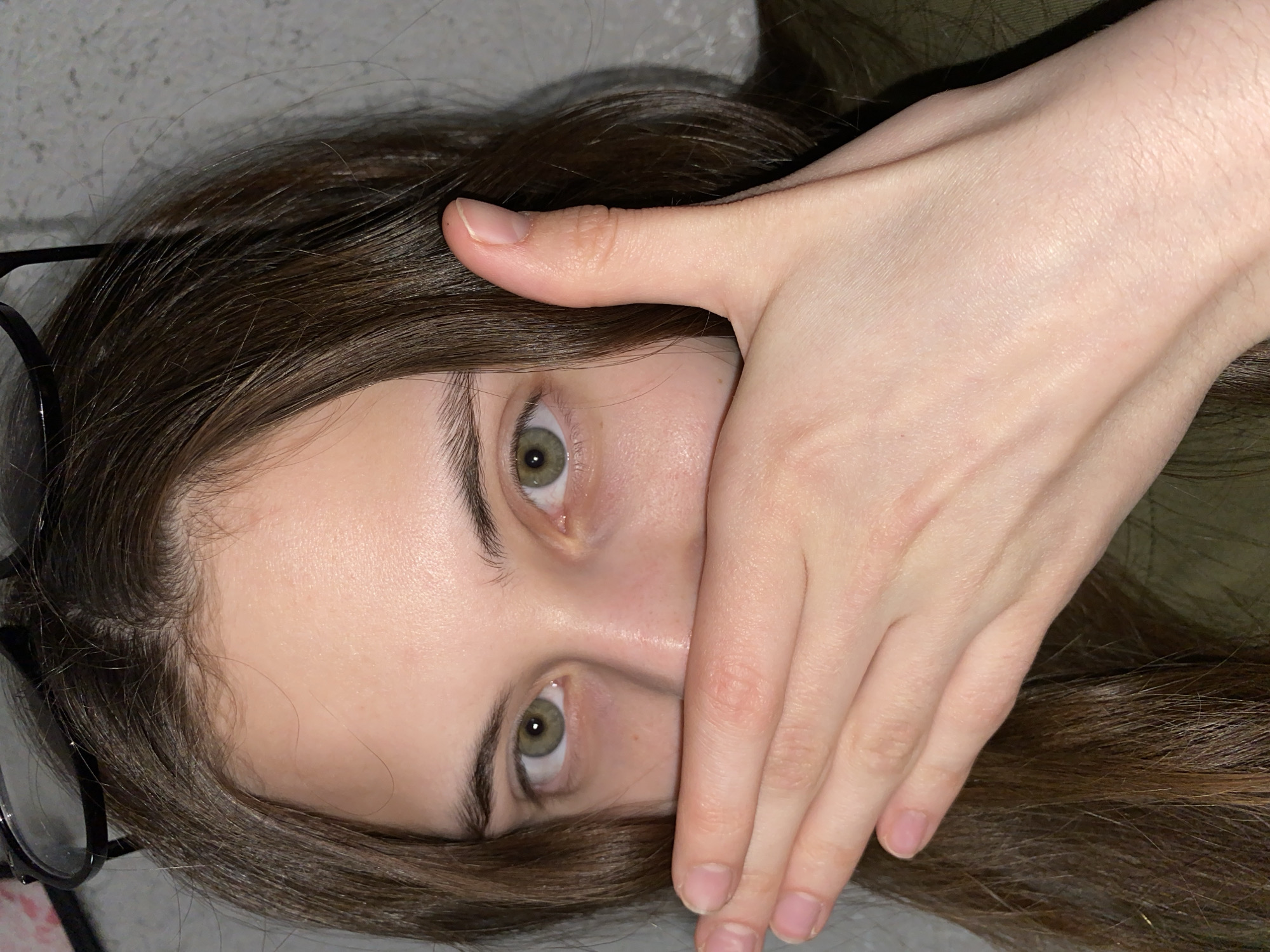 A person is lying down with their hand covering their mouth. The person has brown hair and green eyes. Glasses are visible next to the person's head. The background appears to be a light gray surface. There is no information to determine the image location is Votkinsk, Russia.