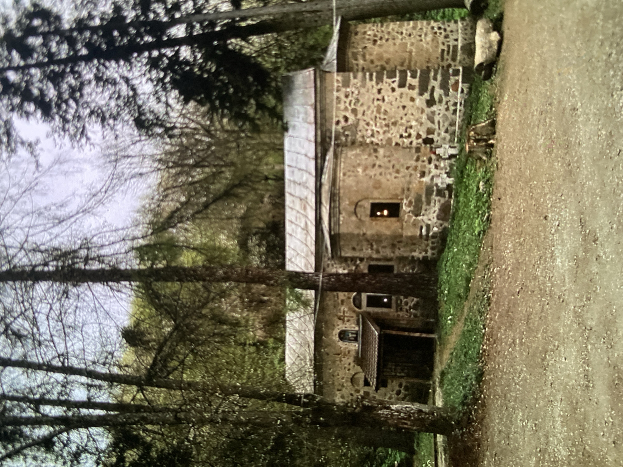 The image shows a rustic stone building nestled among trees, with a dirt path running alongside it. The building, possibly a small church or monastery, is constructed from irregular stones in varying shades of brown and grey, held together with dark mortar. Its roof appears to be made of weathered, light-colored material, possibly metal or slate, with distinct sections.
A prominent feature of the building is a small, arched window on an upper level, from which a soft, warm, yellowish light emanates, suggesting interior illumination like a candle or lamp. This light hints at either dusk, late afternoon, or early morning. A lower entrance or covered porch with a dark opening is visible towards the front of the building.
Tall trees frame the scene, with bare-branched deciduous trees on the left and denser, darker foliage, possibly coniferous, in the background and behind the building. The sky appears light and overcast or hazy. A wide, well-worn dirt or gravel path occupies the foreground and right side of the image, bordered by a strip of green grass along the building's stone wall.
No people or specific activities are visible, contributing to a tranquil and serene atmosphere. The scene evokes a sense of timelessness and seclusion, indicative of a historical site in a natural setting, consistent with its location context of Bankya, Bulgaria. No visible text is present in the image.