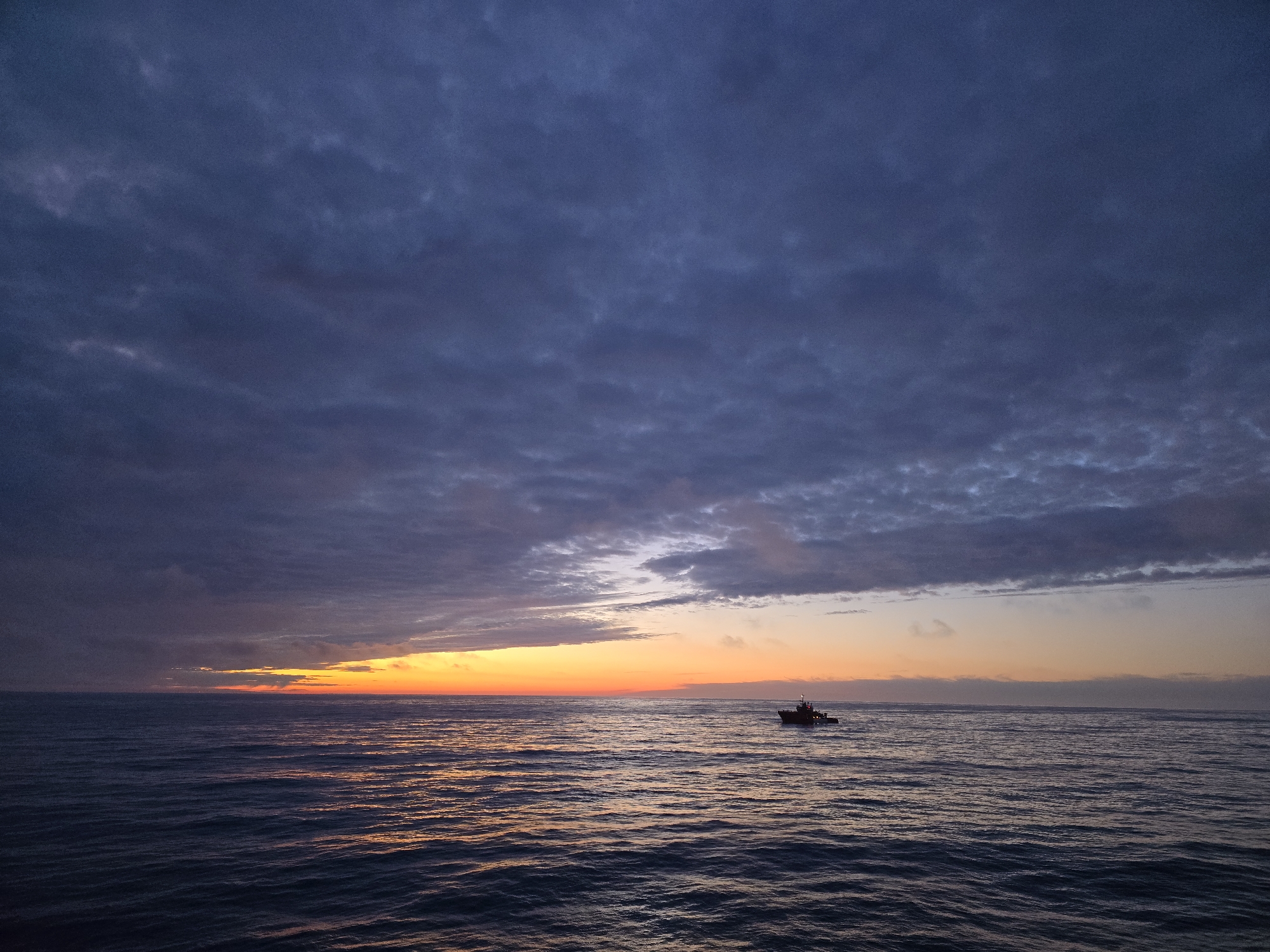The image depicts a body of water, likely an ocean, under a dramatic sky dominated by dark, textured clouds. A sliver of orange and yellow light indicates the presence of a setting or rising sun on the horizon. A small boat is visible on the water's surface. The location is Unknown, Unknown.