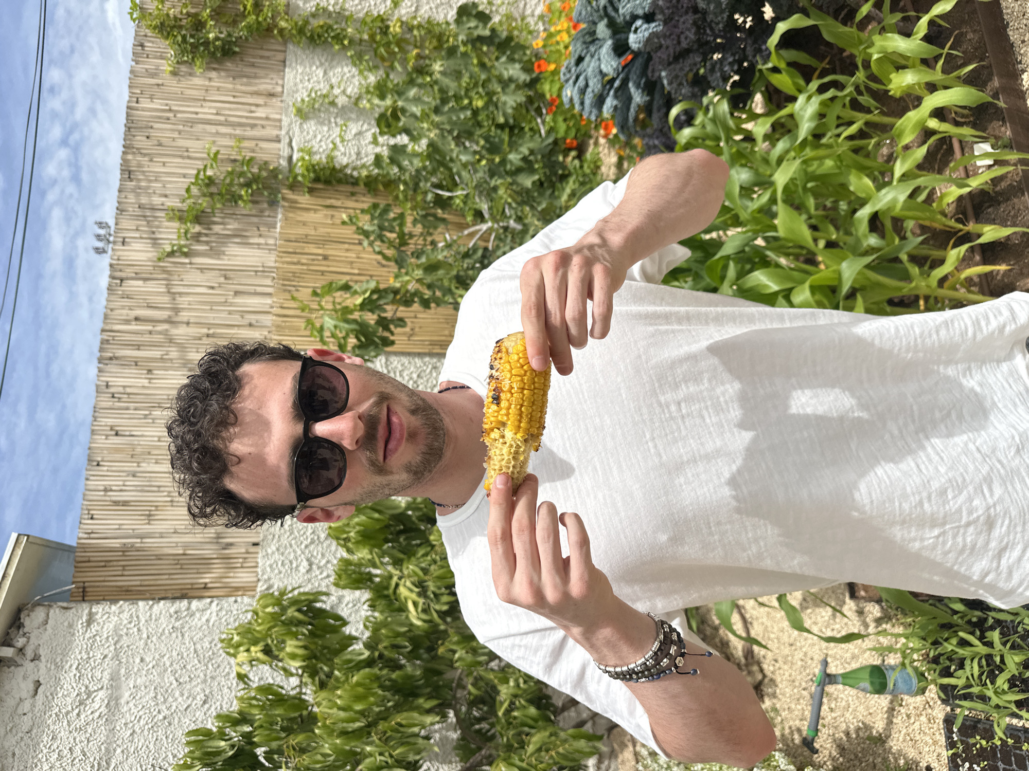 A man with curly dark hair and sunglasses is holding and eating grilled corn in a garden. The background includes bamboo fencing, green foliage, kale, and other plants. A portion of a white building is visible on the left side of the frame. A partially consumed green glass bottle and gardening tools can be seen on the ground. It is daytime with a partly cloudy sky visible in the background. There is no specific information indicating the location is in Tel Aviv, Israel.