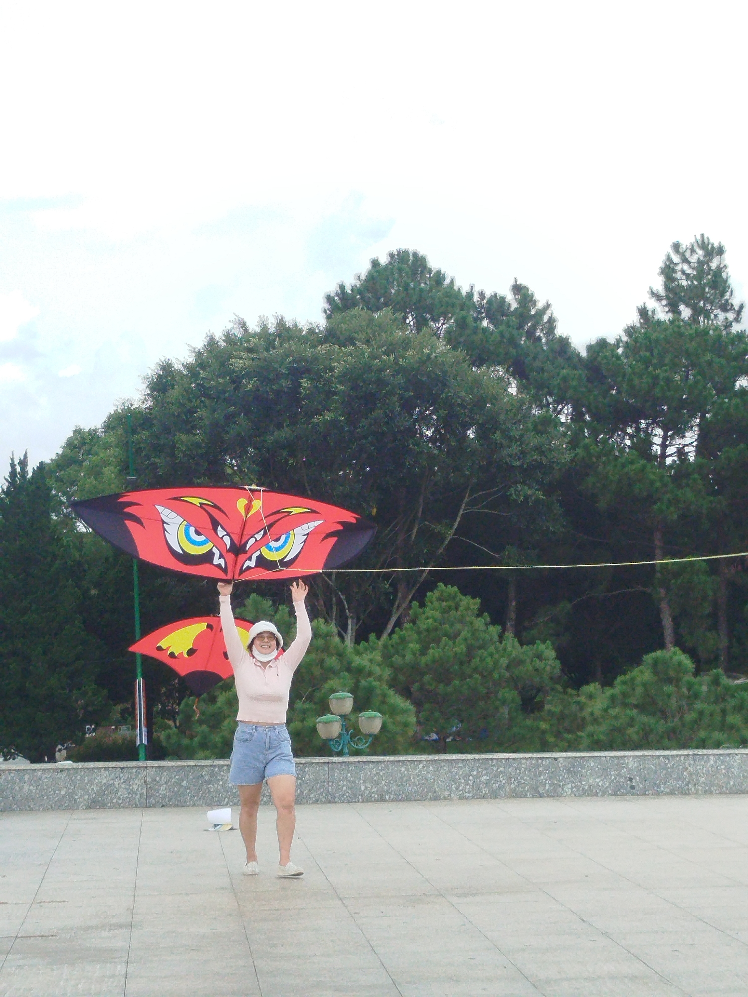 In Da Lat, Vietnam, a young woman stands on a paved plaza, holding a large, red kite shaped like an owl's face above her head. The kite features striking yellow and blue eyes and black accents. She wears a light pink long-sleeved shirt, denim shorts, a white hat, and white sneakers. Another kite, partially visible behind her, is also red with yellow accents. Behind the plaza, a low, textured stone wall separates the paved area from a dense, green forest of pine trees and other foliage. The sky above is overcast, with a bright white hue. The woman appears to be enjoying a day outdoors.
