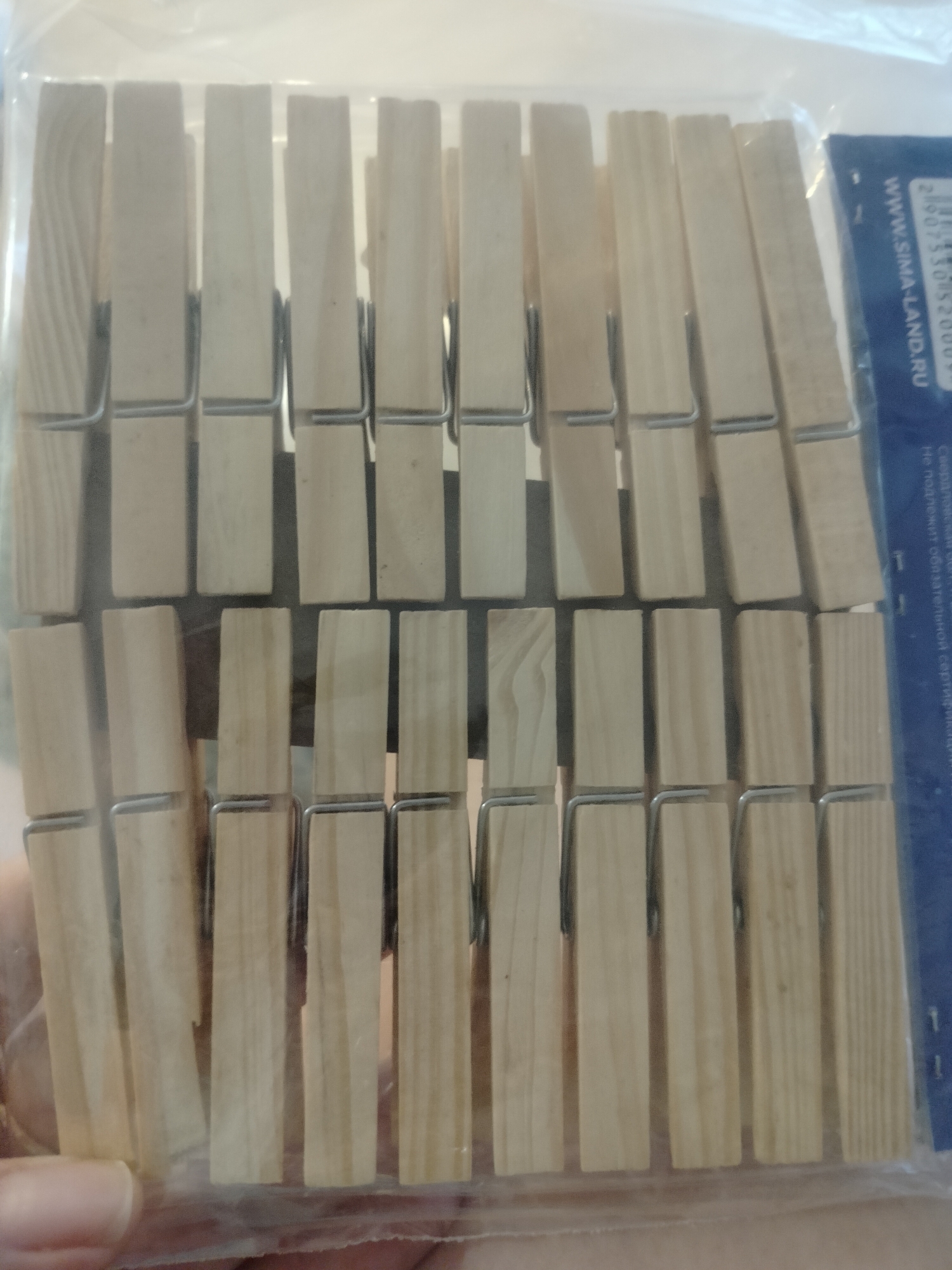 The image shows a package of wooden clothespins. The clothespins are arranged in rows and columns within a clear plastic wrapper. A portion of a blue label with white text is visible on the right side of the image. The text on the label appears to be a website address, "WWW.SIMA-LAND.RU", and a barcode. The background is a light-colored, out-of-focus surface. The lighting suggests an indoor environment, possibly a store shelf or a home setting. There is no discernible information about the location in Novosibirsk, Russia.
