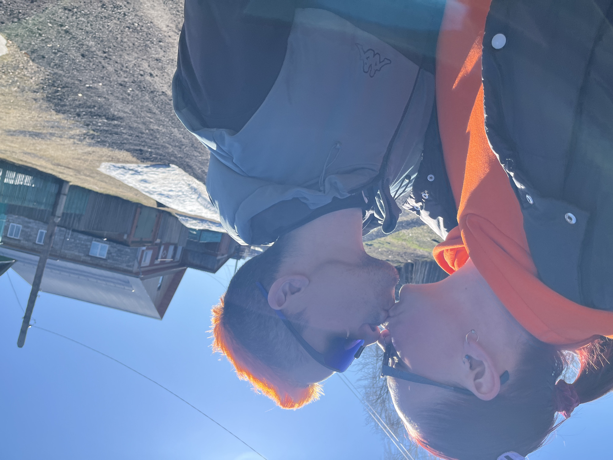 A photograph depicts two individuals in close proximity, potentially kissing, positioned outdoors. The person on the left has short hair dyed orange, wears sunglasses, and is dressed in a Kappa branded vest over a dark garment. The individual on the right has longer, darker hair with pink highlights, ear piercings, sunglasses, and is wearing an orange hoodie with a dark jacket.
The background includes a building and street, indicating an urban or semi-urban environment, with remnants of snow on the ground. This is consistent with the reported location of Chaikovsky, Russia. The angle of the photograph is rotated. The sky is visible and is light blue. A utility pole and wires are also visible.