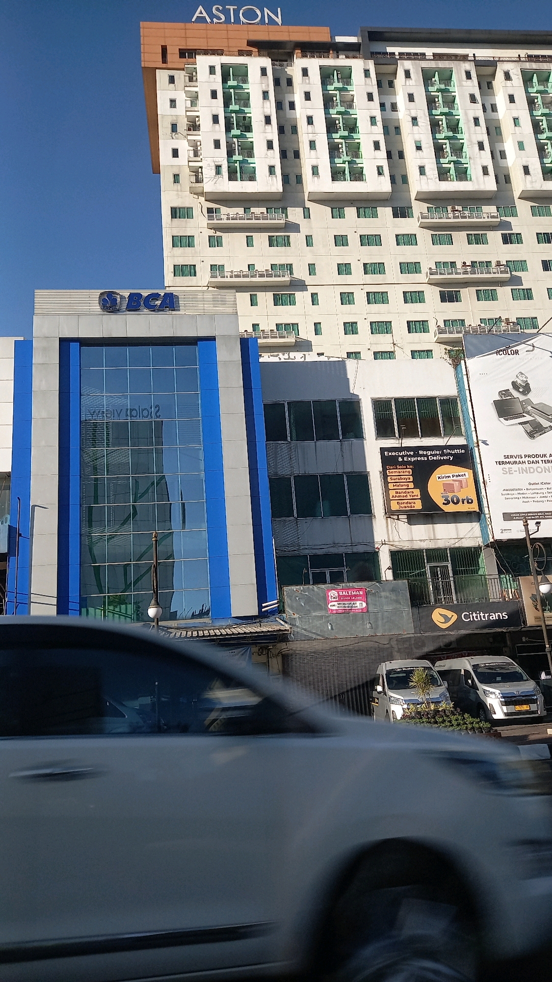 A daytime, eye-level, wide shot captures the Aston Hotel and a BCA building in Surakarta, Indonesia. The Aston Hotel, a tall, beige structure with white and green accents, dominates the background. In the foreground, a modern, blue and white BCA building features a large glass facade. A white car is blurred in the foreground, moving past the buildings. Commercial signage, including "Cititrans" and a package delivery advertisement, is visible on a lower building situated between the BCA building and the foreground car. The sky is clear and blue.