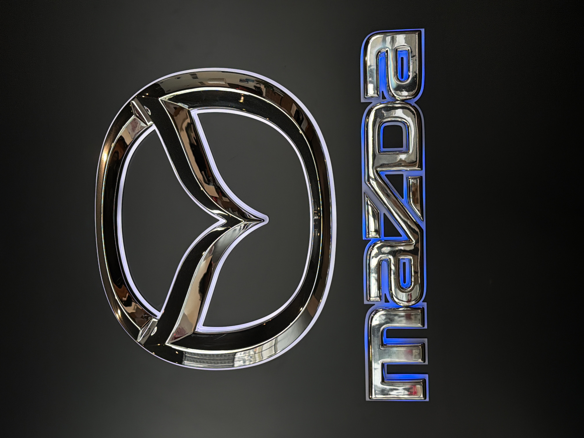 The image shows the Mazda logo and name against a black background. The logo, a stylized "M" inside a circle, is silver with a reflective surface. The name "Mazda" is displayed vertically in silver lettering with blue accents, also featuring a reflective surface. There is no information in the image to suggest a location in the city of Izhevsk, Russia or any people or actions.