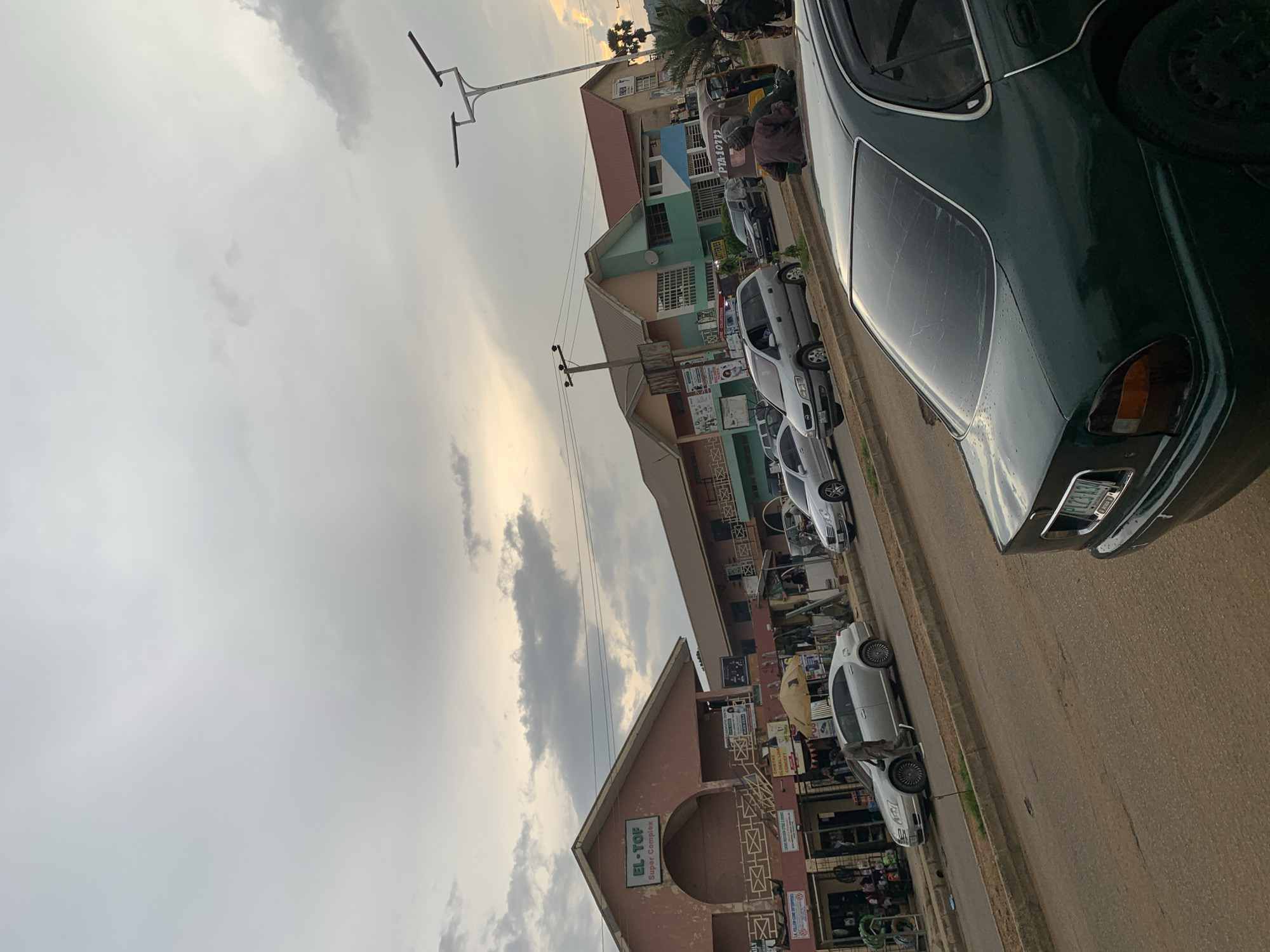 The image depicts a street scene in Rafin Par, Nigeria, characterized by a cloudy sky. Multiple buildings line the right side of the street, including one prominently labeled "EL-TOF Super Complex." Cars, a tricycle, and pedestrians are present on the asphalt road. Several vehicles are parked or in motion along the street. The buildings appear to be commercial establishments, with storefronts visible. The overall atmosphere suggests a typical urban environment with moderate activity.