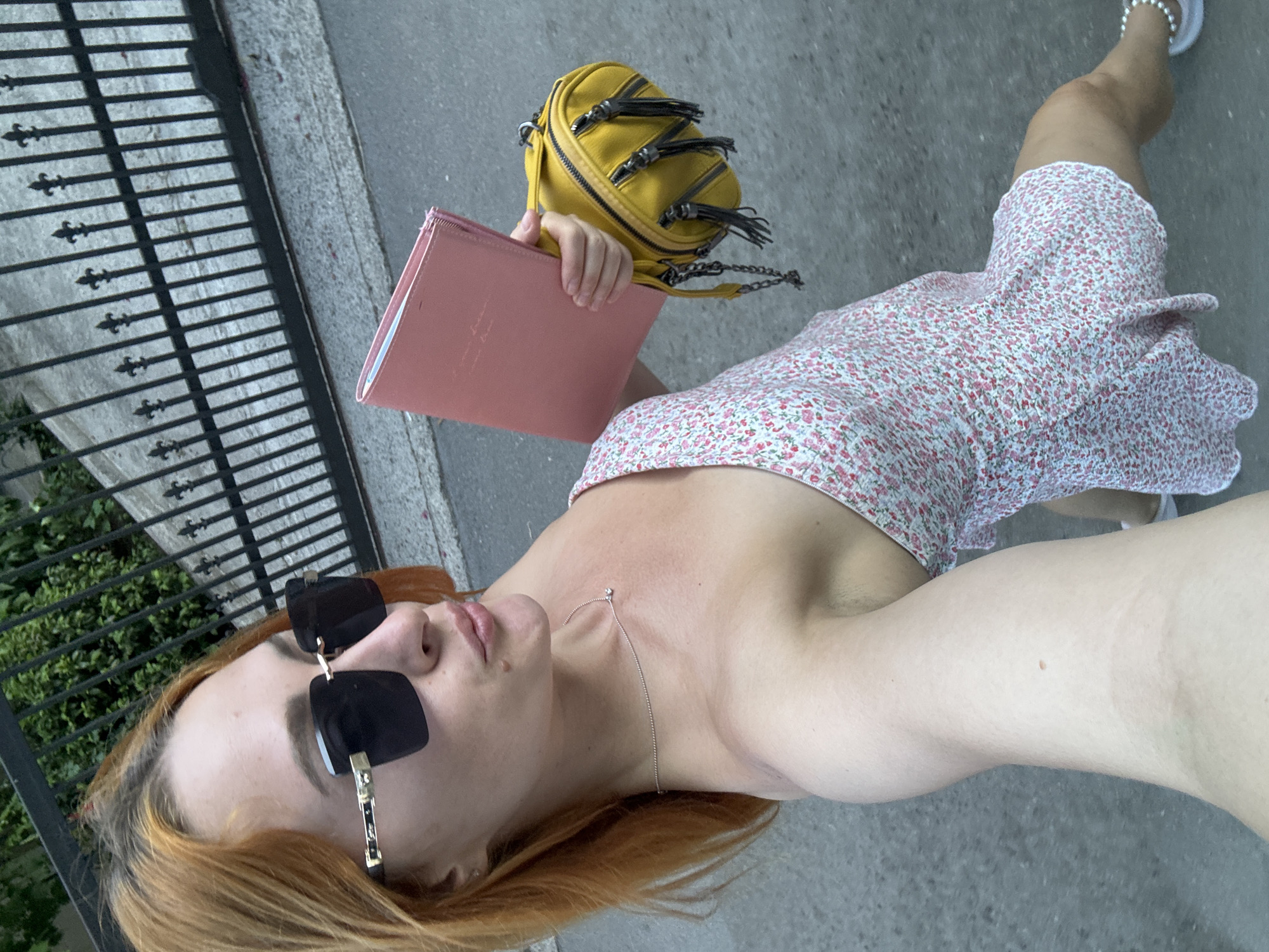 A woman with reddish-blonde hair, wearing large dark sunglasses, a delicate silver necklace, and a short floral-print dress, is standing on a paved surface. She is holding a pink notebook in her left hand and a small yellow crossbody bag with fringe detailing in her right. Her lips are slightly puckered. Behind her, a black metal fence with decorative finials separates the paved area from a patch of green foliage. The scene appears to be outdoors, possibly on a street or sidewalk, with the architecture and vegetation suggestive of a European city like Budva, Montenegro.