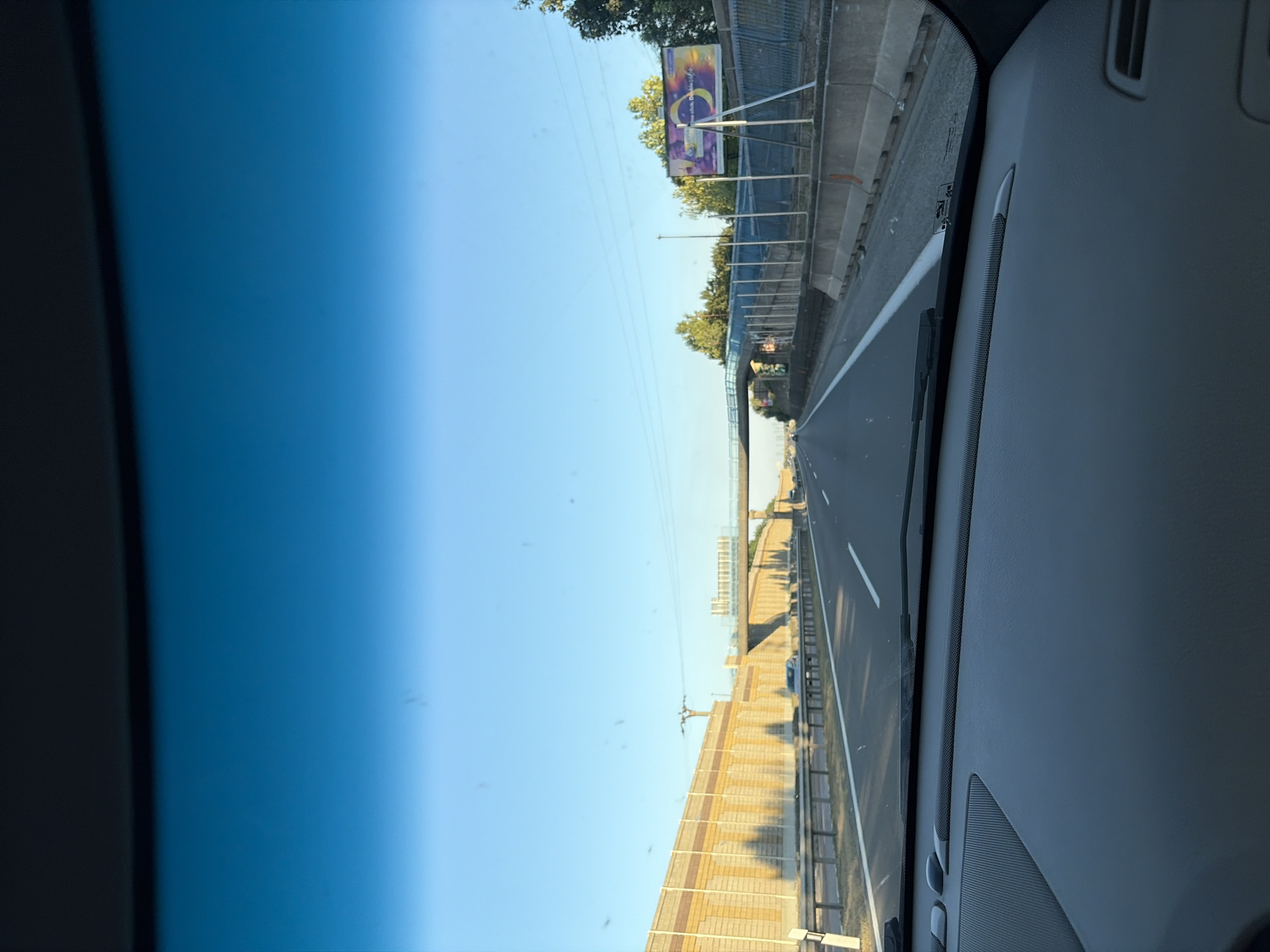 The image shows a view from the inside of a car driving on a highway. The highway has multiple lanes and is flanked by a sound barrier on the left and trees and a billboard on the right. In the distance, a pedestrian overpass crosses the highway. The sky is clear and blue. The dashboard of the car is visible in the foreground. The location is Vlčkovce, Slovakia.