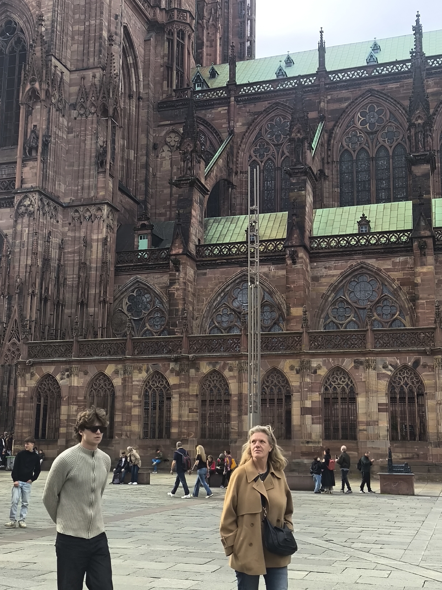 The image depicts a public square in Strasbourg, France, with the Strasbourg Cathedral visible in the background. The architecture of the cathedral is characterized by intricate Gothic details and red sandstone construction. Several people are scattered throughout the square, engaged in various activities. In the foreground, a young man wearing sunglasses and a zip-up sweater walks toward the left. To his right, a woman in a tan coat and jeans stands looking up. Other people are standing, walking, or sitting in the background. The ground of the square is paved with stone blocks. The sky appears overcast, suggesting diffused lighting.