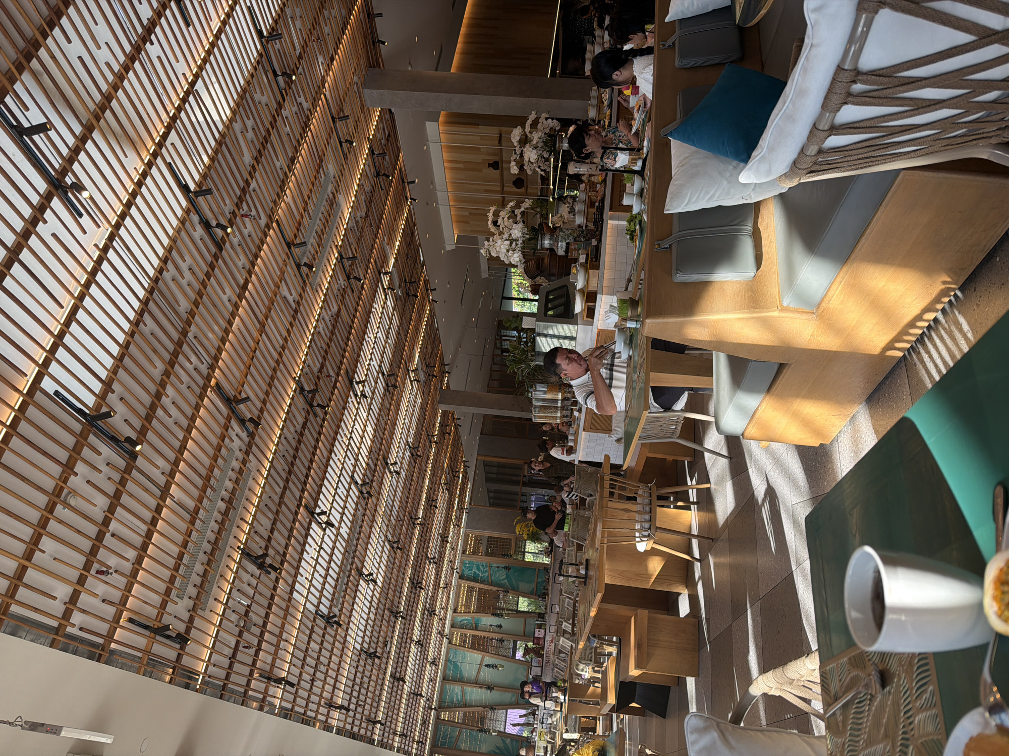 The image depicts a brightly lit indoor restaurant with a distinctive wooden slatted ceiling and modern lighting fixtures. Sunlight streams in, casting geometric shadows on the tiled floor, suggesting a location with ample natural light. The interior is furnished with light-colored wooden tables and chairs, as well as cushioned seating areas.
Several patrons are visible in the restaurant, engaged in dining and socializing. In the foreground, a white mug with a dark liquid sits on a green table. In the midground, a man in a striped shirt is eating at a table, and a woman with dark hair is seated next to him. Further back, other diners are spread across the various seating arrangements. The restaurant appears to be a buffet or a dining hall with multiple serving stations visible in the background, adorned with floral arrangements and food displays. The overall ambiance is relaxed and contemporary, fitting for a mealtime setting in a modern establishment. The restaurant is located in Dong Ninh Hoa Ward, Ninh Hoa Town, Ninh Hoa City, Khanh Hoa Province, Vietnam.