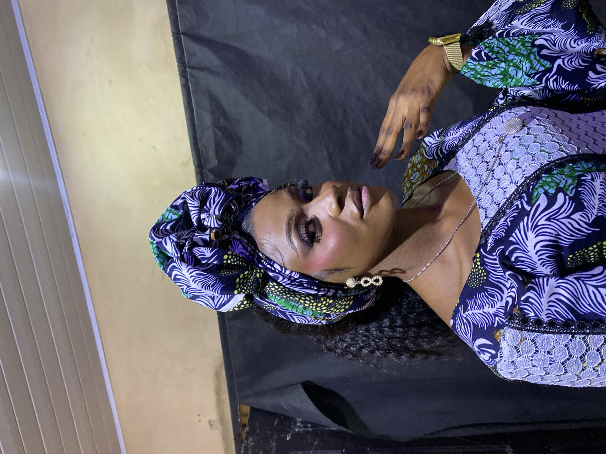 A portrait-style image features a woman with dark skin, elegantly posed. She is looking upwards and slightly to the right, towards the camera. She wears a vibrant, multi-patterned headwrap, predominantly blue, green, and yellow. Her attire appears to be a dress or top, combining a similar blue and white leaf-like or feathered pattern with sections of light purple or white lace.
Her accessories include a gold-colored watch with a mesh band on her left wrist, a thin silver chain necklace with a small, round pendant, and earrings that resemble an infinity symbol, possibly adorned with small gems. Her dark, curly hair is visible around her face. Her makeup is pronounced, with defined eyebrows, dark eye makeup, and light-colored lips. Her fingernails are long, manicured, and painted with a dark polish. One hand is raised near her head in a graceful gesture.
The background is divided, with a light beige or yellowish wall on the left side and a dark, possibly black fabric or curtain, covering the right side and behind the subject. A vertical structure, possibly blinds or panels, is partially visible on the far left. The lighting is artificial, highlighting the subject's features and intricate outfit. The scene appears to be an indoor setting for a posed photograph. There is no visible text in the image.