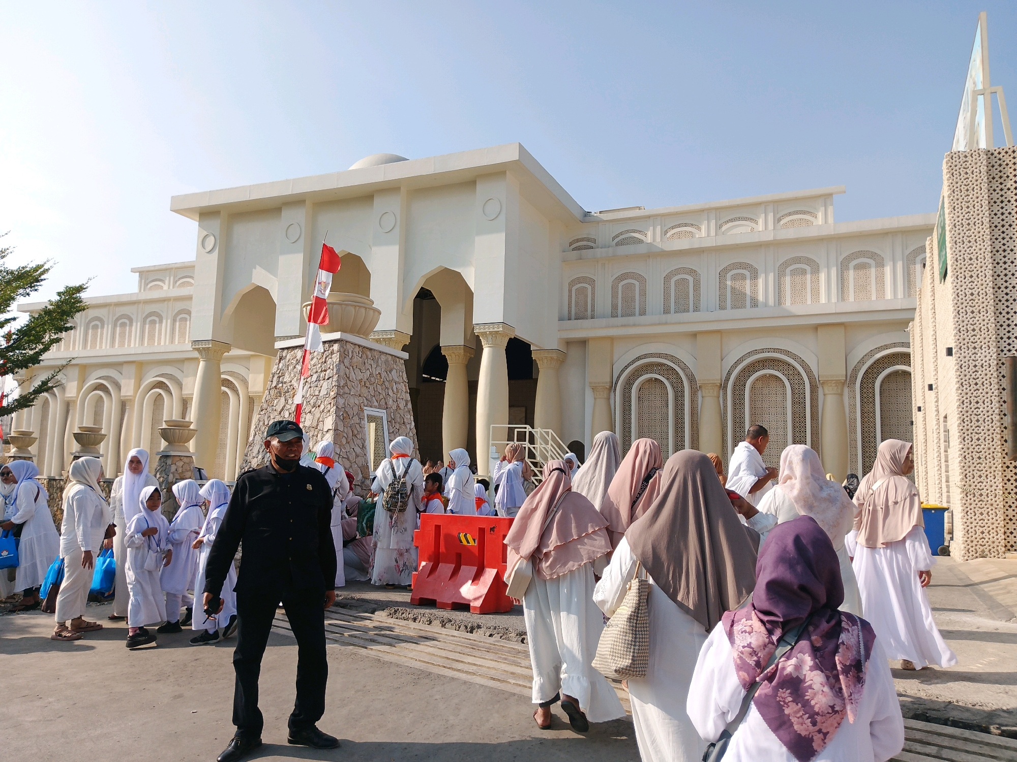 A man in a black uniform and a black cap stands in the foreground of an outdoor scene, facing left. He wears a mask. Behind him, a group of people, predominantly women and children, are walking and standing, many wearing white clothing and hijabs. The building behind them is a large, modern structure with intricate architectural details, including arches and decorative patterns. An Indonesian flag flies on a pole near the entrance. The scene appears to be a public gathering or event.