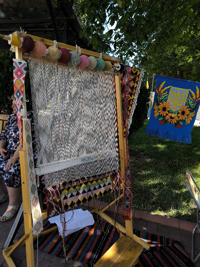 The image depicts an outdoor weaving exhibit or demonstration in Illintsi, Ukraine, illuminated by bright daylight. Central to the scene is a tall, light-brown wooden weaving loom with numerous white warp threads stretched vertically. Nine colorful skeins of yarn, ranging from maroon to black, are suspended from the top crossbar of the loom. A section of a partially woven textile, displaying horizontal geometric patterns in black, pink, yellow, blue, red, and green, is visible near the base of the warp threads. Two narrow, vertically oriented textiles with traditional geometric patterns are attached to the side supports of the loom.
To the right of the loom, a rectangular blue textile banner is suspended, featuring a central yellow Ukrainian Trident emblem surrounded by a wreath of yellow sunflowers and red berries, with a fringed bottom edge. Below the loom, a striped rug with horizontal bands of black, red, blue, and green is laid on a light-colored paved surface. White paper-like objects are situated on this rug beneath the loom's weaving area. A small, light-colored wooden stool is positioned on the striped rug to the lower right.
A person is partially visible on the far left, seated on a bench behind the loom. They wear a dark patterned dress and light-colored sandals. The background consists of green grass, bordered by a low stone or brick wall, and lush green tree foliage in the upper and upper-right portions of the frame. A dark overhead structure with a translucent roof is visible at the top, providing partial shade to the setup.