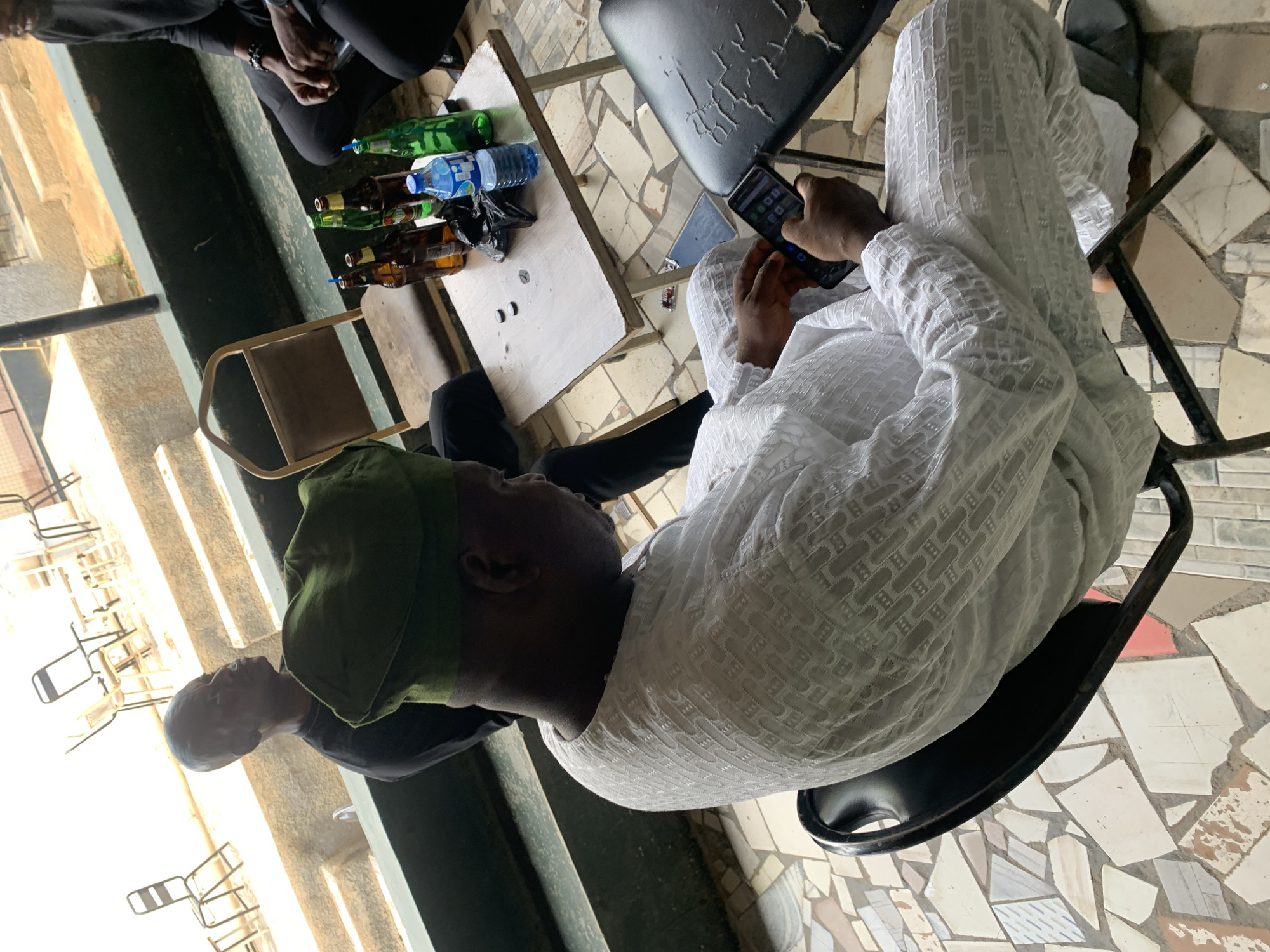 This image depicts a casual outdoor gathering in Kabong, Nigeria, under natural daylight. In the foreground, a man with dark skin is seated on a black chair, observed from a high-angle perspective. He wears a white, long-sleeved traditional garment with a distinct textured pattern and an olive green cap. He is actively interacting with a dark-colored smartphone, held in both hands, displaying various app icons on its screen. Dark sandals or slides are visible on his feet.
To the immediate left of the central figure, a second dark-skinned man is partially visible, wearing a dark shirt and looking towards the upper right side of the frame. To the right of the scene, the lower body and hands of a third individual, dressed in dark attire, are visible near a small, light-colored rectangular folding table with metal legs.
The table contains several items: at least four brown glass beer bottles, two green glass bottles (one fitted with a blue straw), two plastic water bottles (one with a blue label), a small black plastic bag, and two small dark circular objects. An empty light brown chair with a metal frame is positioned adjacent to the table. The ground is covered with an irregular mosaic pattern of light-colored tiles, including shades of white, beige, and grey, with occasional reddish fragments. Behind the seating area, a dark green ledge or barrier separates the foreground from a lower level featuring concrete steps and a lighter-colored ground surface. In the background, multiple empty metal-framed chairs and small tables are visible on an elevated, light-toned platform.