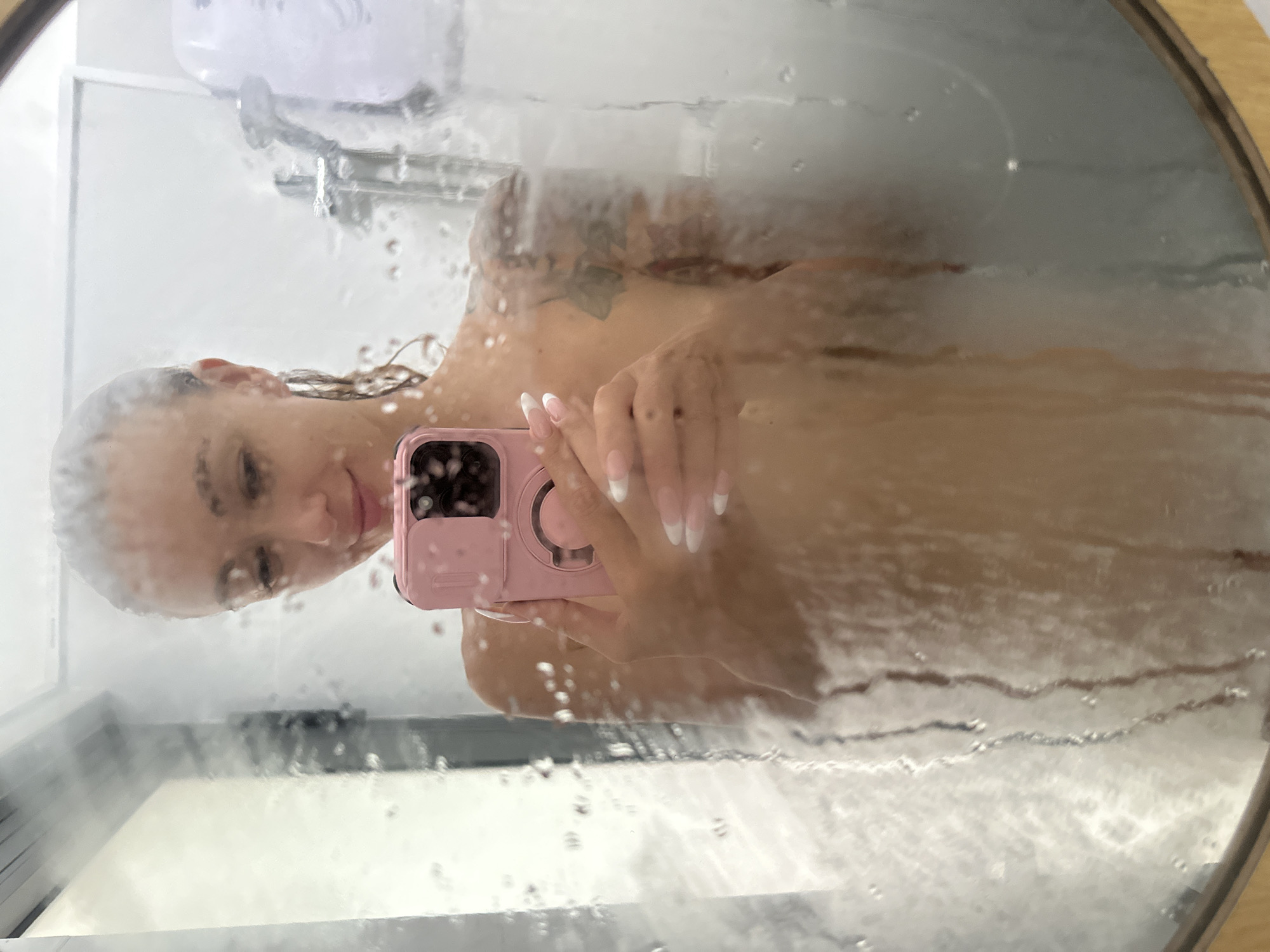 The image captures a close-up selfie of a young woman reflected in a round or oval mirror, heavily covered in condensation and water droplets. The setting appears to be a bathroom or shower in Mũi Né, Vietnam, indicated by the steamy mirror and visible shower fixtures in the blurred background.
The woman is seen from the chest up, with her head tilted and her gaze directed towards the smartphone she holds. Her hair is wet and slicked back, and she wears a soft, relaxed expression with a slight smile. Notable details of her appearance include a tattoo, possibly floral or nature-themed, visible on her right shoulder and neck. Her hands, holding the phone, display long, manicured fingernails with a distinct French tip design.
She is holding a smartphone encased in a prominent pink protective case, featuring a visible camera module and a ring stand or pop socket on the back. The surface of the mirror is textured with numerous water droplets and streaks, which blur and diffuse the reflection, lending an artistic, soft-focus effect to the scene. The background behind her reflection shows light-colored walls and vague outlines of a shower head or faucet. There is no visible text in the image.