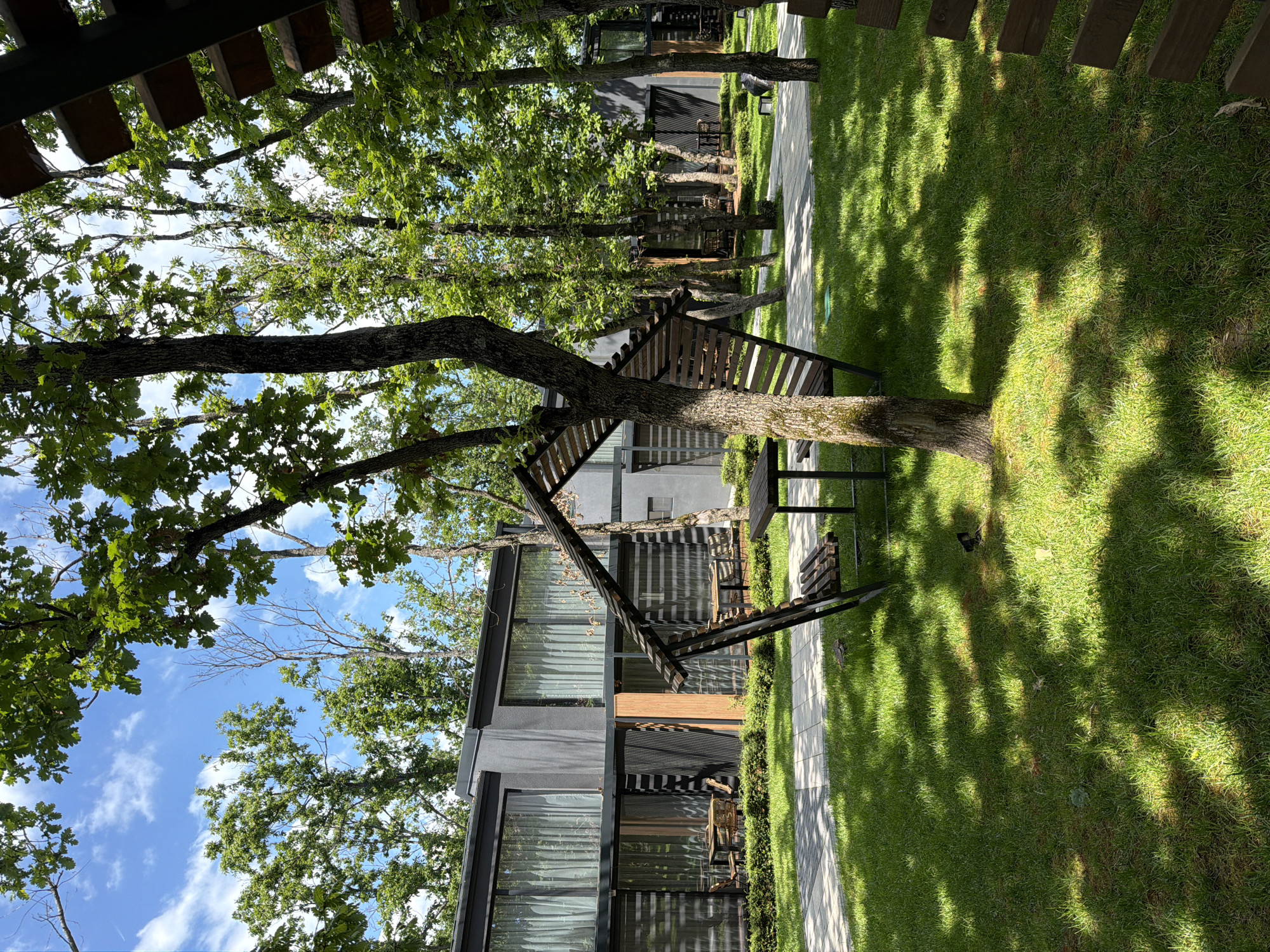 The image depicts a modern building with large windows, partially obscured by trees. A wooden structure resembling an outdoor staircase or decorative feature is attached to a tree in front of the building. A sidewalk runs alongside a well-maintained grassy area, with shadows cast by trees adding texture. The scene appears to be in an urban or suburban environment, possibly a park or recreational area. Based on the prompt's specification, this might be Burgas, Bulgaria, but this cannot be definitively confirmed from the image itself. There are no people visible in the frame.