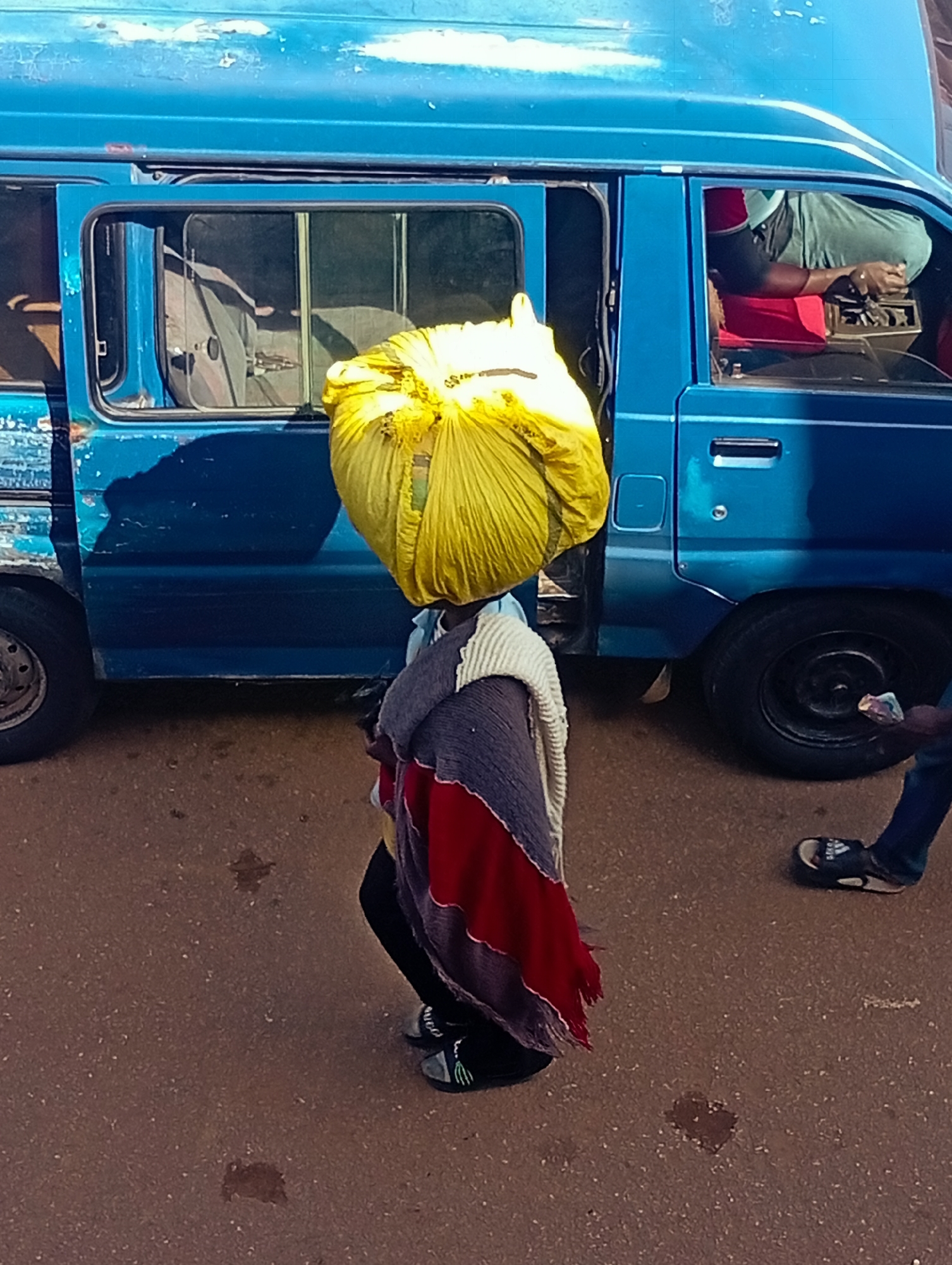 In Bukuru, Nigeria, a person walks beside a blue van with a large yellow bag balanced on their head. The person is wearing a multi-colored shawl draped over their shoulders and a dark garment underneath. Their feet are adorned with black sandals. The van, appearing weathered with chipped paint, has its side door ajar, revealing part of the interior. A driver is visible in the driver's seat, hands on the steering wheel. The ground is paved with reddish-brown asphalt, marked with scattered small dark spots. The background shows the side of the van and a portion of the road.