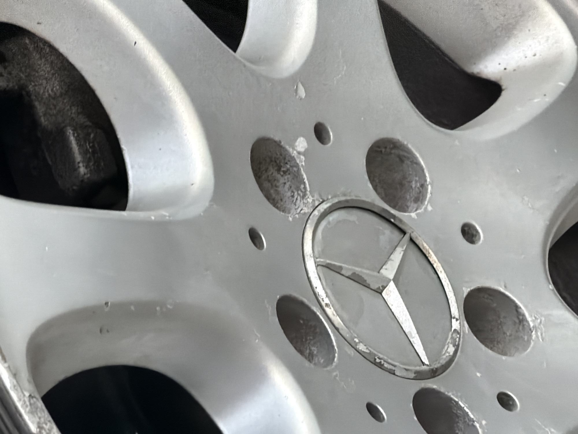 The image shows a close-up of a silver Mercedes-Benz car wheel, likely in Plovdiv, Bulgaria. The wheel exhibits signs of wear and tear, with some corrosion or pitting visible within the lug nut holes and around the center cap. The Mercedes-Benz logo is present in the center of the wheel. The background is dark and out of focus, suggesting the car is parked or in a shaded area. There are no people or other objects clearly visible in the image.
