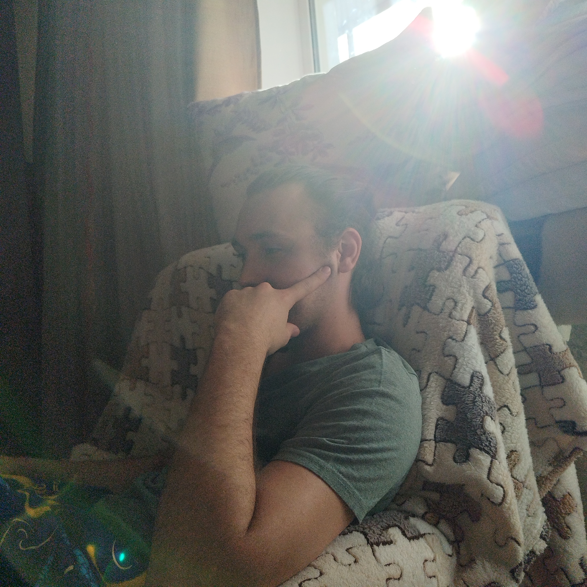 A Caucasian man is resting in an armchair near a window. The location is most likely Yekaterinburg, Russia. The man is wearing a green t-shirt and has his left arm raised with his index finger touching his left cheek. He appears to be in thought or contemplation. The armchair is covered with a blanket patterned with puzzle pieces. A window is visible behind the armchair, with sunlight streaming through. Dark curtains are to the left of the armchair.
