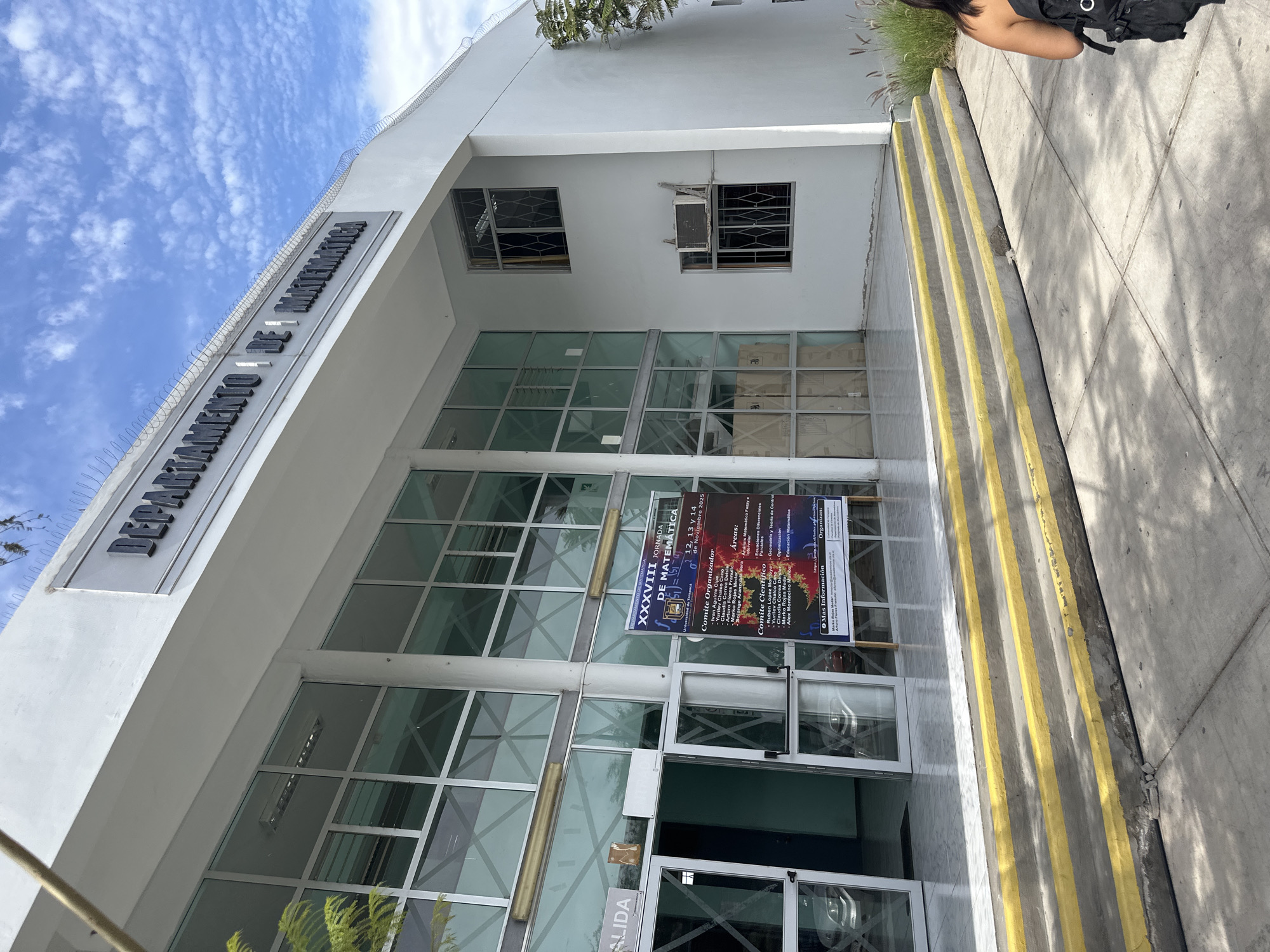 The image shows the entrance to the Departamento de Matemática (Department of Mathematics) at the Universidad de Tarapacá in Arica, Chile. A sign on the building identifies the department. The building is modern with a white exterior and large glass windows and doors. A poster advertising the XXXVIII Jornada de Matemática (38th Mathematics Conference), scheduled for November 12-14, 2025, is displayed on the front doors. The poster lists the organizing committee members and the areas of mathematics to be covered. A flight of stairs with yellow markings is located to the right of the entrance. A person with a black bag is partially visible at the top of the stairs. The sky is blue with light clouds. Barbed wire is visible on the top of the building.
