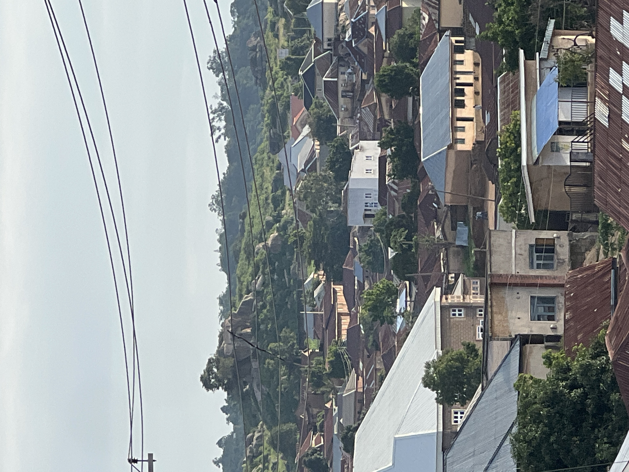 The image depicts a dense residential area in the city of Jos, Nigeria, with a hilly, vegetated landscape in the background. The foreground is dominated by various buildings with corrugated metal roofs, characteristic of many urban and semi-urban areas in Nigeria. Several structures feature multi-colored roofs, including rusted brown, gray, and light blue. The architecture varies, with some buildings appearing to be single-story homes, while others are larger, multi-story residential complexes. Prominent among these is a white, modern-looking building with multiple windows on the upper floor.
The middle ground shows a cluster of buildings with terracotta and brown tiled roofs, interspersed with greenery. Power lines stretch across the upper portion of the image, suggesting an active utility infrastructure. The background features rolling hills covered in lush green vegetation and scattered rock formations, indicating the natural topography of the area surrounding Jos. The sky is overcast and light blue, suggesting a daytime scene, possibly in the late morning or afternoon. The overall impression is one of a vibrant, lived-in urban environment integrated with its natural surroundings. No people are clearly visible in the image.