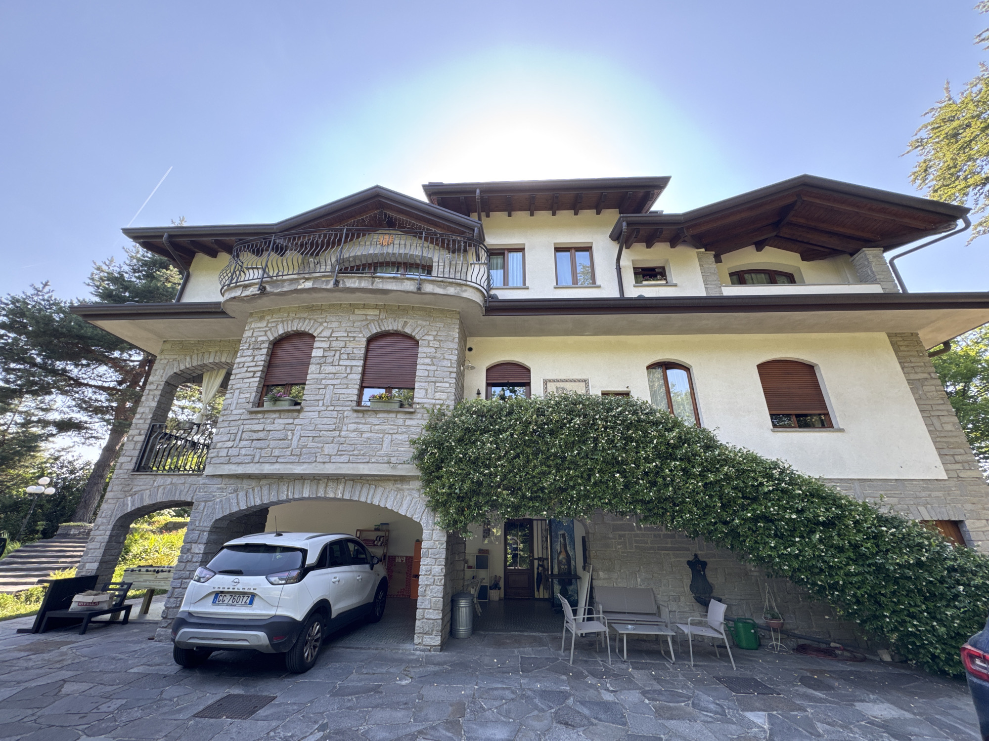 A wide-angle, low-angle exterior shot captures a large, multi-story house in Cunardo, Italy, on a clear, sunny day. The house features a light-colored facade with stone accents around windows and doors. A prominent arched driveway is visible, with a white Opel Crossland SUV parked within it. Lush green ivy cascades over the side of the house, partially obscuring the entrance. A wrought-iron balcony extends from the upper level, and several windows with brown shutters are visible. In the foreground, a stone patio is laid out, with outdoor furniture including chairs and a small table. To the left, a foosball table and benches are set up, and a set of stone stairs leads up the side of the property. The sky is a bright blue with a faint airplane contrail. The overall scene depicts a well-maintained residential property with a strong emphasis on natural elements and outdoor living.
