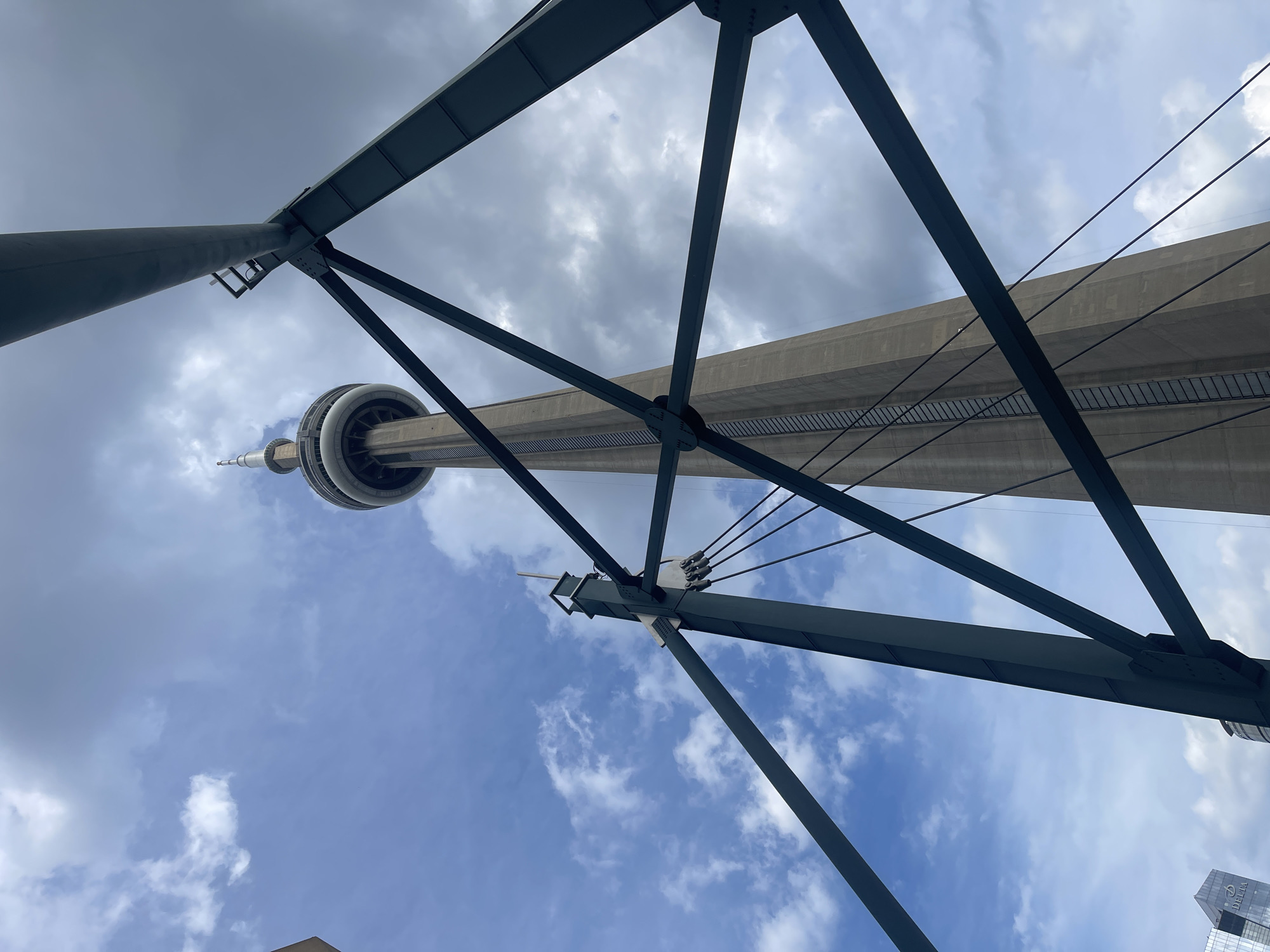 This image captures a low-angle view of the CN Tower in Toronto, Canada, framed by the dark, angular metal structure of what appears to be a bridge or overpass. The iconic tower, a towering concrete structure with a distinctive observation deck and spire, rises prominently against a partly cloudy sky. The metal framework in the foreground, with its diagonal beams and cables, creates a geometric contrast with the organic shapes of the clouds and the smooth, vertical lines of the tower. The lighting suggests it is daytime, with a mix of sunlight and shadow playing on the structures and clouds. In the lower left corner, a sliver of a modern glass building with a visible logo is present, indicating an urban setting. There are no people or animals visible in the frame.