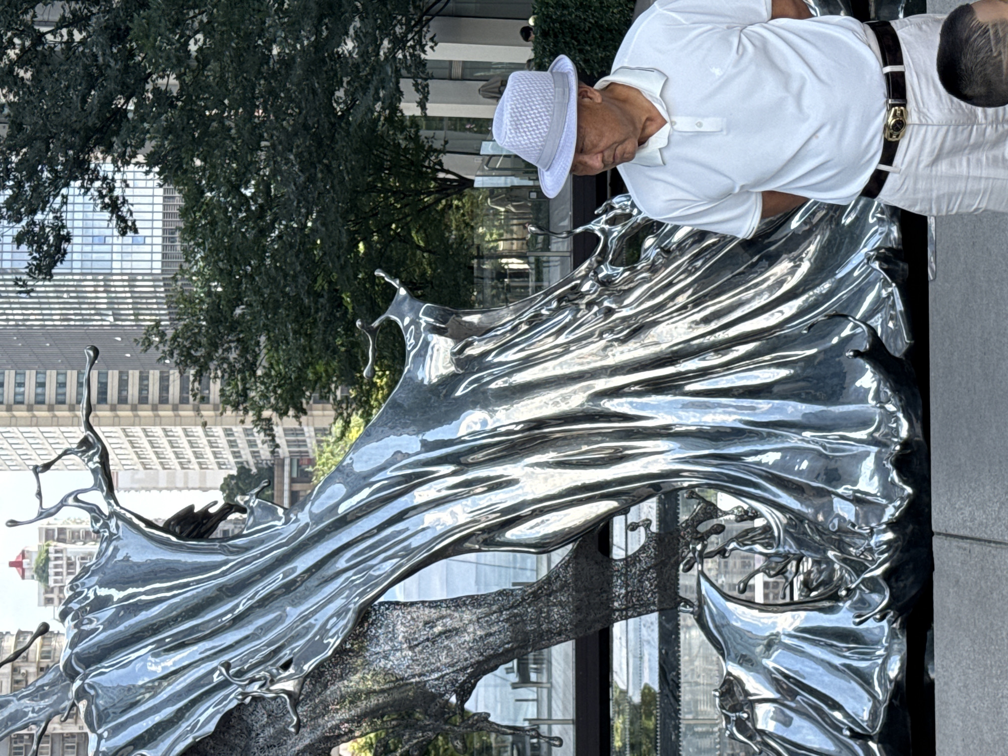 In Yuzhong District, China, a man wearing a white polo shirt, white pants, and a light-colored hat stands to the right of a large, abstract, silver sculpture. The sculpture has a fluid, wave-like appearance with sharp, metallic edges. Behind the sculpture, a body of water is visible, reflecting the metallic sheen of the artwork. In the background, modern buildings with glass facades rise into the sky, interspersed with the dark green foliage of trees. The overall scene appears to be an urban outdoor setting, possibly a public plaza or park, with a focus on the interplay between art, nature, and architecture.
