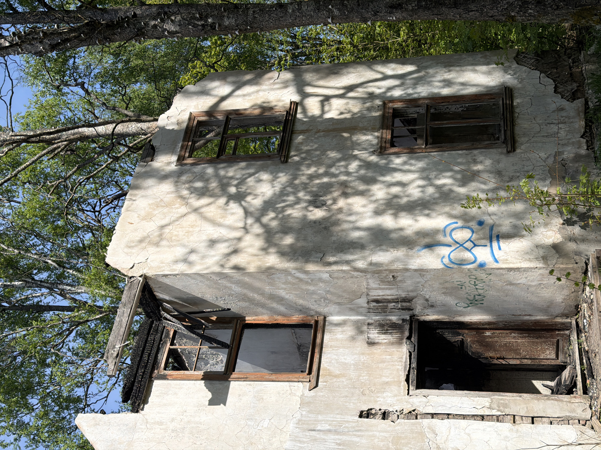 The image shows a dilapidated, partially destroyed two-story building in Tokkari, Russia. The structure has a crumbling white facade with visible cracking and damage. Four window openings are visible. The top two are empty with wooden frames. The bottom two also have wooden frames; one is damaged by fire, the other is completely burnt out. The building's facade is marked with blue graffiti. Shadows of trees cover the building's exterior, and trees surround the building itself. The overall environment suggests neglect and abandonment.