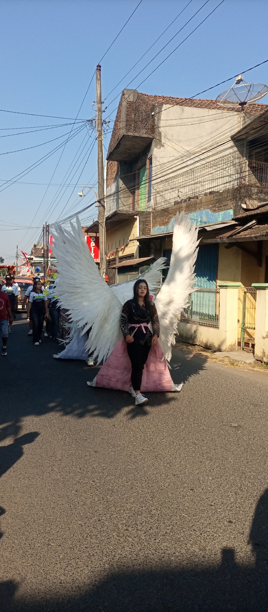 In the city of Jekulo, Indonesia, a young woman is captured in a frontal, medium shot, standing in the middle of a paved street. She is adorned with large, white, feathered wings, reminiscent of an angel's costume. The wings are spread out behind her, with the tips extending outwards. The woman wears a dark patterned top with a pink sash tied around her waist, black pants, and white sneakers. Her gaze is directed forward, with a neutral expression. A pink-and-white layered fabric drapes from her waist down to the ground, extending past her feet. In the background, several people are walking in a procession, some wearing matching t-shirts. Buildings with weathered facades and visible utility poles and wires line the street, suggesting an urban or semi-urban environment. The sky is clear and blue, indicating a sunny day. The image is taken from a slightly low angle, emphasizing the scale of the wings. Shadows of people are visible on the asphalt in the foreground.