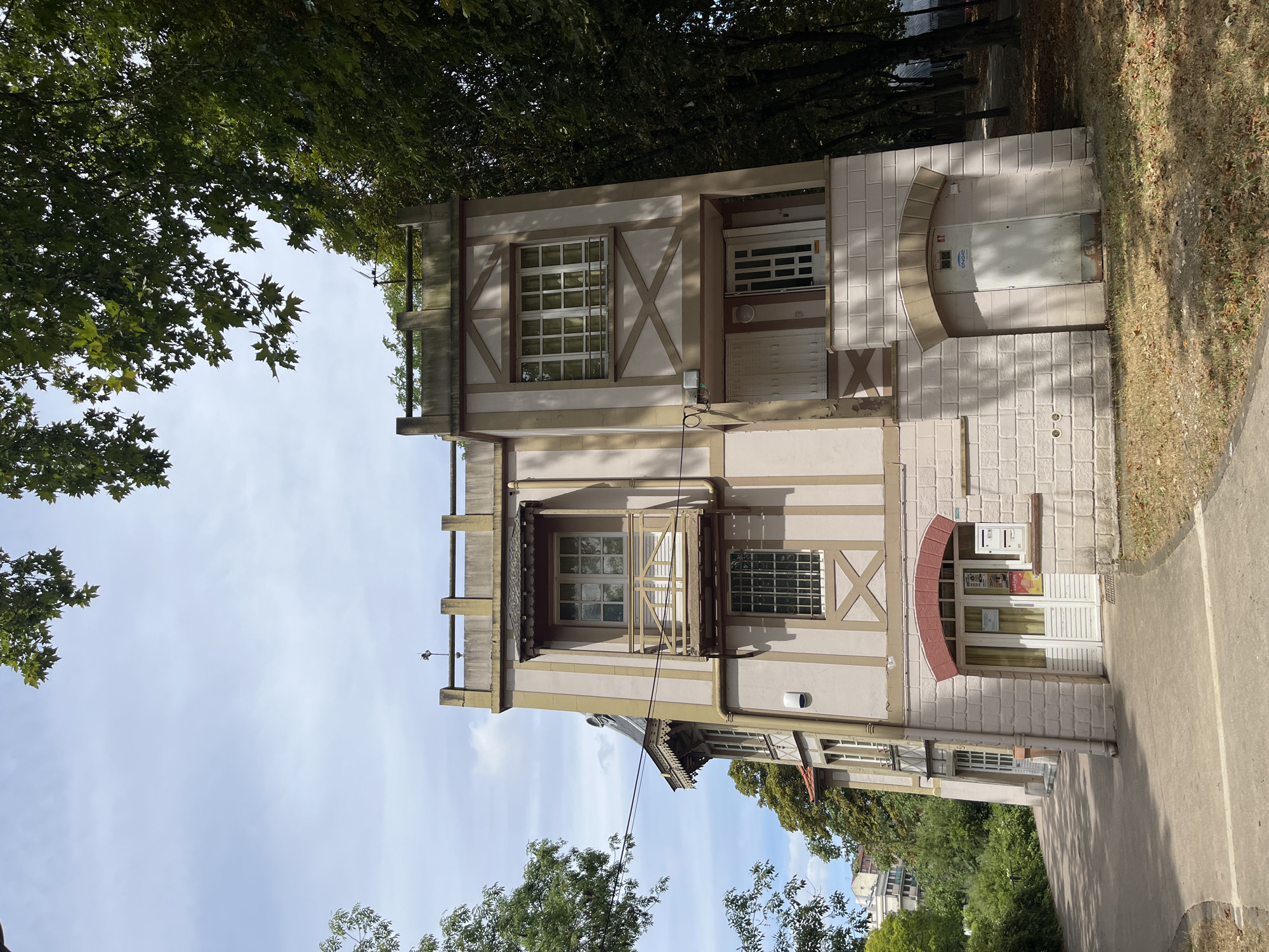 The image shows a three-story building with light pink and beige facade, located in Joinville-le-Pont, France. The building is situated on the right side of the frame, with a pathway and vegetation on the left. The building has several windows, some with wooden balconies and awnings. The ground floor features two doors, one with a red brick archway and the other a white door under a beige archway. A mailbox is visible to the right of the door with the red brick archway. Trees with green foliage surround the building, with a path leading away from the structure into a wooded area. The sky is partly cloudy.