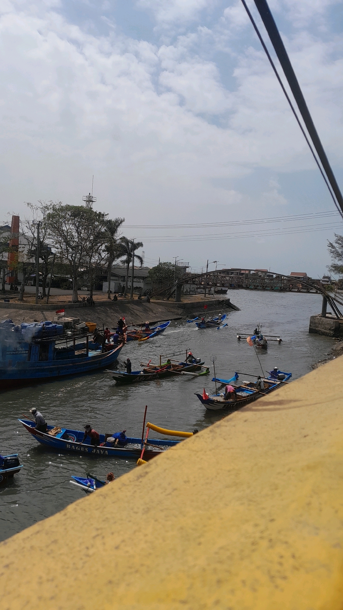 The image depicts a waterway in Kelurahan Tegalkamulyan, Cilacap, Indonesia, filled with various boats. In the foreground, a yellow railing partially obscures the view. A large blue boat, labeled "BAGUS JAYA," is prominently featured, with several individuals on board, some appearing to be working. Several smaller fishing boats, with outriggers and often adorned with small flags, are scattered throughout the waterway. These boats are occupied by people, many of whom are dressed casually and appear to be engaged in activities related to boating or fishing. In the background, a curved pedestrian bridge spans the waterway, with buildings and trees lining the banks. The sky is partly cloudy. The overall scene suggests a busy waterfront with ongoing maritime activity.