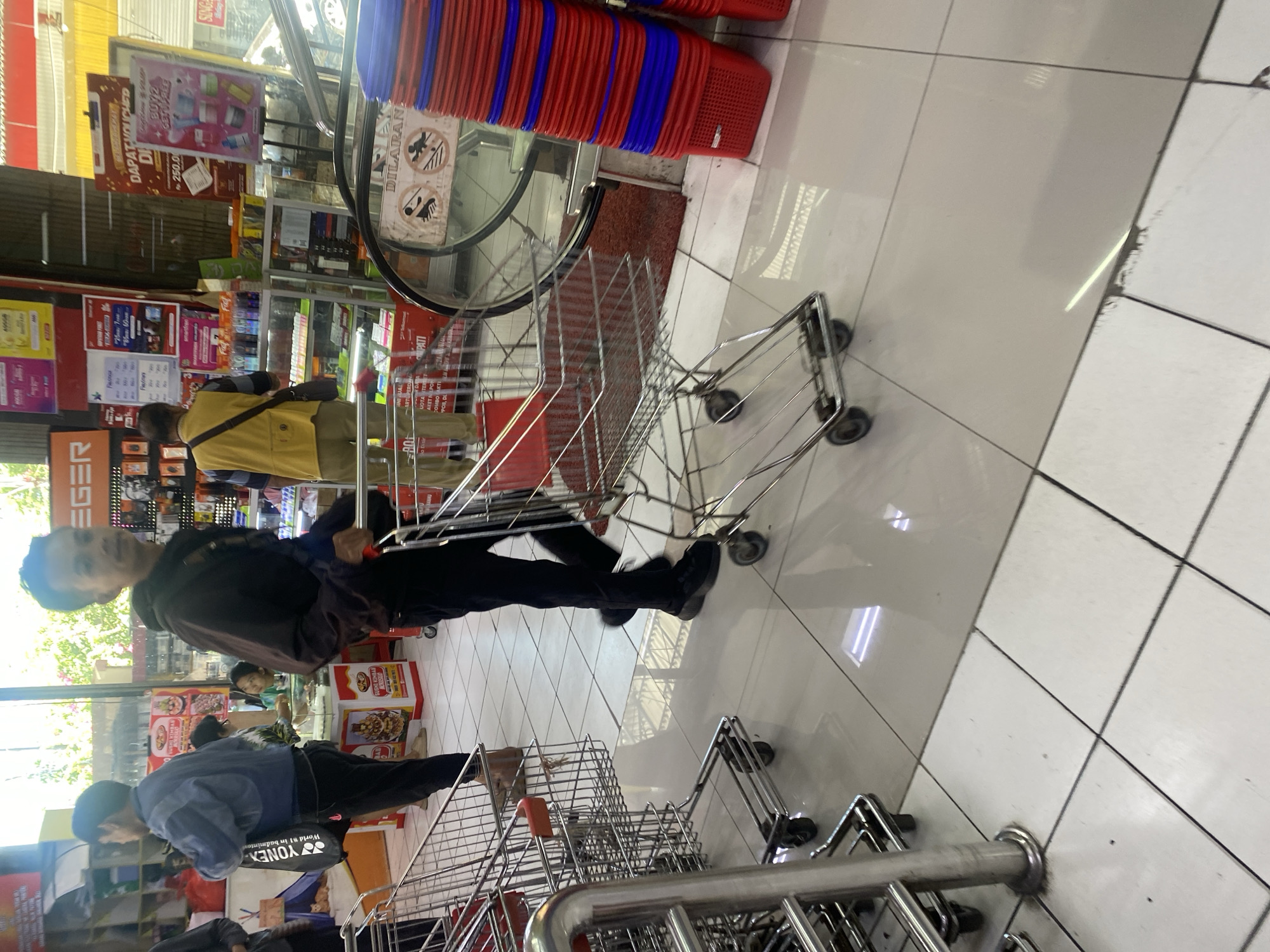 The image depicts the interior of a supermarket, likely in Manado, Indonesia, as indicated by contextual clues like signage and typical store layout. The scene is busy with several shoppers and a variety of merchandise.
In the foreground, a man in dark pants and a dark hooded jacket is pushing a metal shopping cart. He is looking towards the camera with a neutral expression. To the left of the frame, another shopping cart is visible, partially obscured by a stainless steel post. Behind the man pushing the cart, another individual wearing a blue jacket is visible, also pushing a shopping cart.
Further in the background, a man in a yellow vest is standing near a display of goods. The shelves are stocked with various products, including what appear to be electronics and toiletries. Red and blue plastic baskets are stacked neatly to the right of the frame, near an escalator. The floor is tiled in a grid pattern. Natural light enters from a large window or glass entrance, illuminating the store. The overall impression is one of everyday commerce within a retail environment.