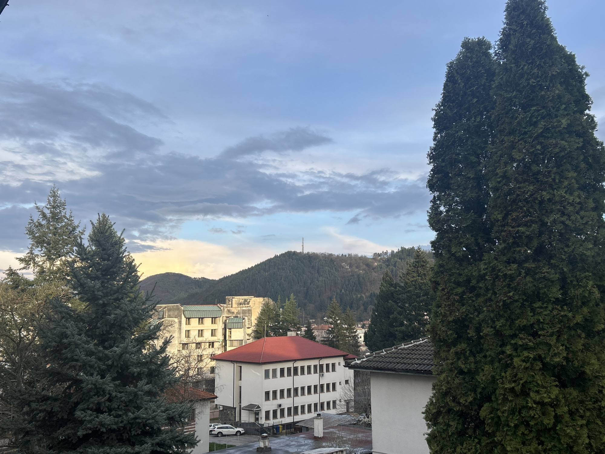 The image shows an elevated view in Zlatograd, Bulgaria. A multi-story building with a red roof is visible in the foreground. Another light-colored building stands behind it. In the midground, a tree-covered mountain rises, with a communication tower on its peak. The sky is partially cloudy with hues of blue and grey. Tall, coniferous trees frame the scene on the left and right sides of the image. A white car is parked near the red-roofed building. The scene appears to be an urban landscape situated near a mountainous region.