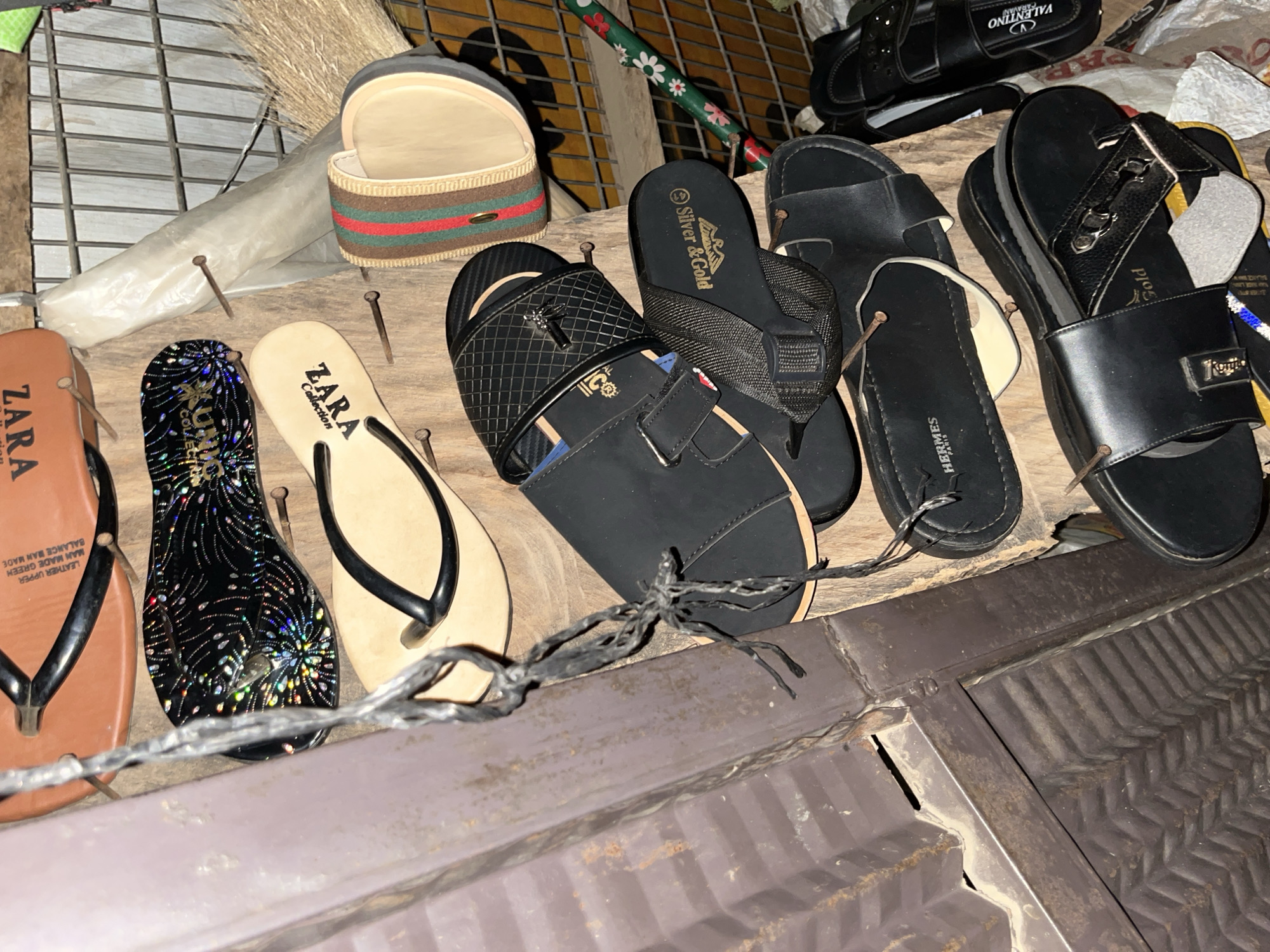 A collection of open-toed footwear, including flip-flops and sandals, are displayed on a wooden surface. The footwear exhibits various designs and brands such as "ZARA", "HERMES", "Silver & Gold", and "VALENTINO". Some items are adorned with decorative elements like glitter and metallic accents. The scene is dimly lit, suggesting it might be evening or a poorly lit area. The background includes a metal grate, a broom, and what appears to be corrugated metal sheeting, all contributing to an outdoor or semi-outdoor market stall aesthetic, possibly located in a commercial area within Abuja, Nigeria. There are no people visible in the image.