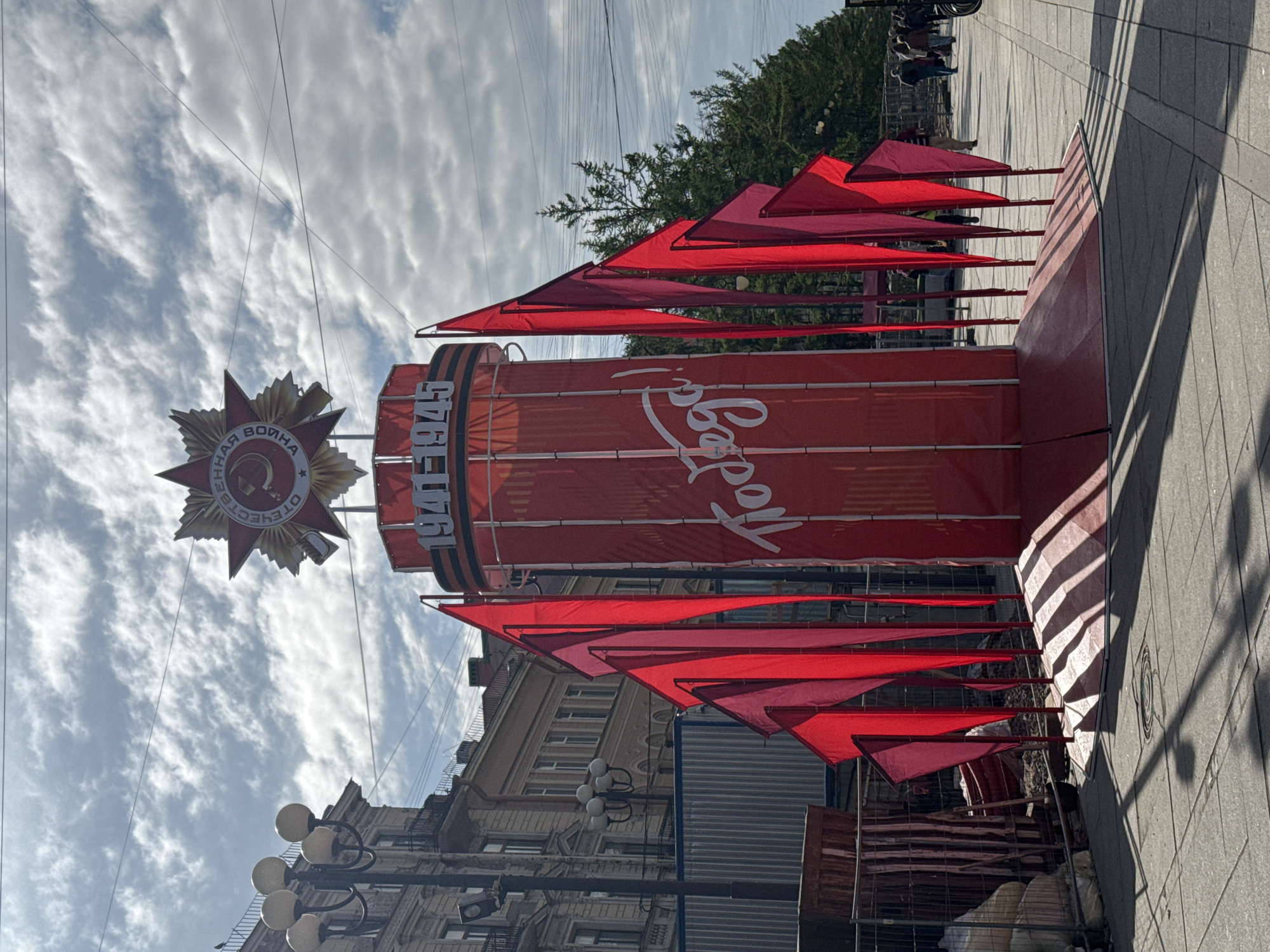 The image shows a red cylindrical structure, possibly for a holiday or event, in Saint Petersburg, Russia. The structure has the number "1945" on it, and the word "Подол" written in script. Above the structure is a red starburst design with Russian text. Red triangular flags are arranged around the perimeter of the structure. The structure is located on a sidewalk in an urban area, with buildings visible in the background, as well as utility poles and wires. There are some people sitting in the background.