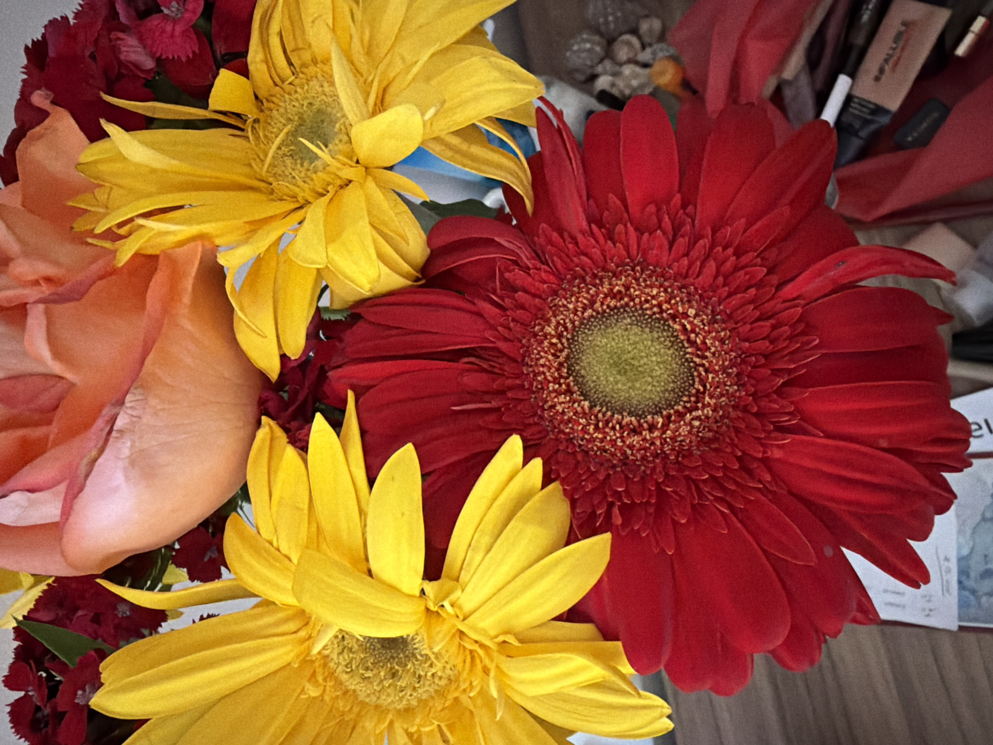 The image shows a close-up of a bouquet of flowers. The bouquet includes two yellow sunflowers, a red gerbera daisy, and a peach rose. The red gerbera daisy is the largest flower in the bouquet and is located on the right side of the image. The two yellow sunflowers are located on the left side of the image, and the peach rose is located on the top left side of the image.
There is no evidence that this image was taken in Catania, Italy.