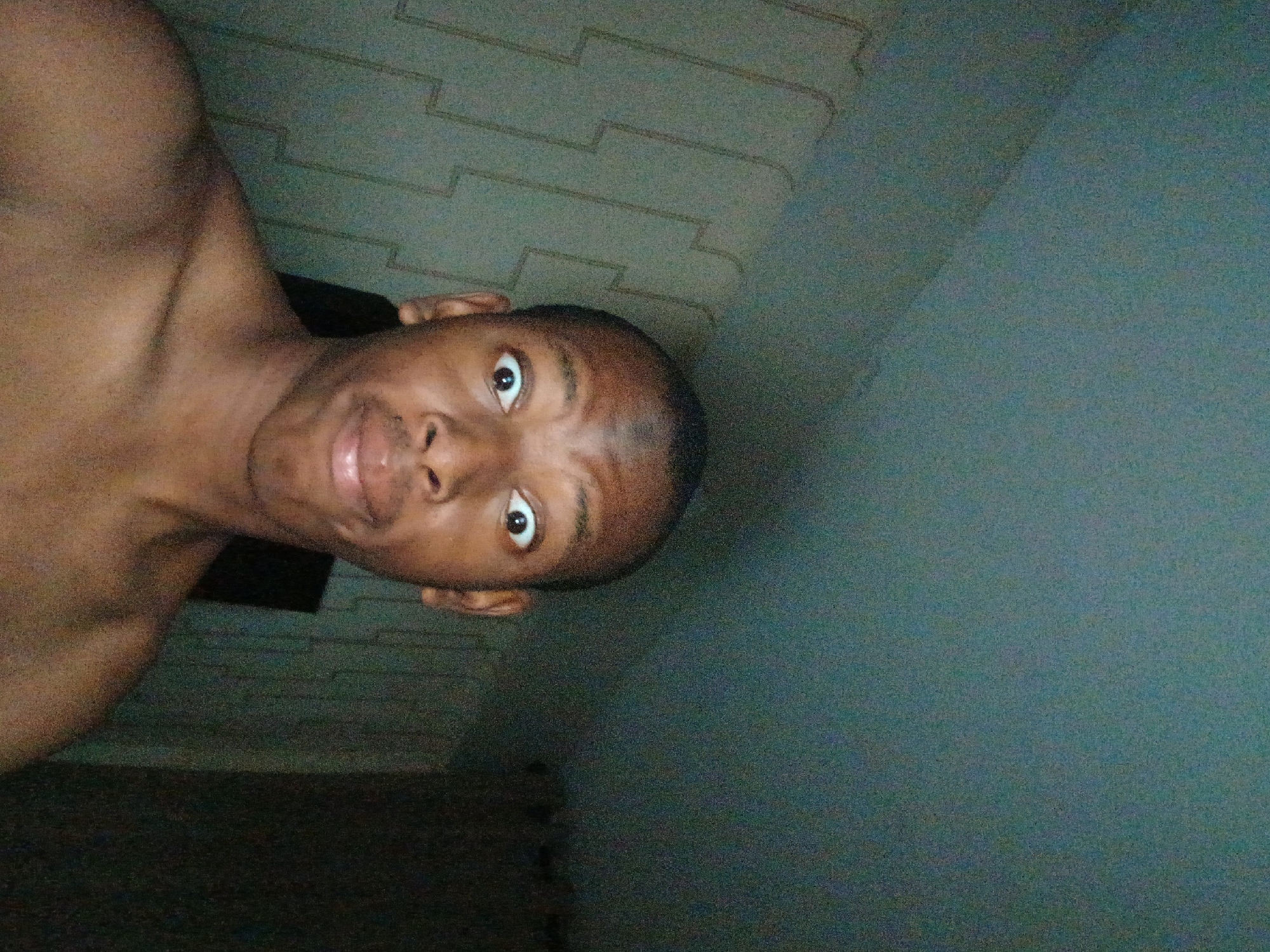 A shirtless young Black man is close to the camera. He has a dark complexion, short hair, and a small amount of facial hair. His eyes are wide open. His mouth is slightly ajar, with his lips slightly puckered. Part of his shoulder is visible.
The background is a mottled, gray-green wall that looks like an indoor setting. A decorative, patterned element of the building runs from left to right.
The image is dark and has a grainy texture, which could indicate it was taken in low-light conditions. The image appears to be taken at an angle.
No specific details provide information regarding the location in the city of Lagos, Nigeria.