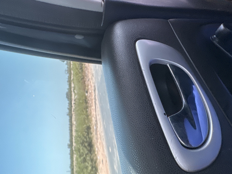 The image depicts a view from the interior of a vehicle in Dokwa, Nigeria, looking out through the side window. The foreground features the upper portion of a car door panel, primarily textured black plastic or faux leather. A chrome-plated interior door handle, set within a silver trim bezel, is centrally located in the lower half of the frame; a blue reflection is prominent on its surface, and minor scratches are visible on the trim. Outside the vehicle, a paved road stretches horizontally across the midground, bordered by a dirt shoulder. Beyond the road, the background consists of an open, vegetated landscape under a clear, bright blue sky, indicating an outdoor, daylight setting. No individuals or specific actions are discernible.