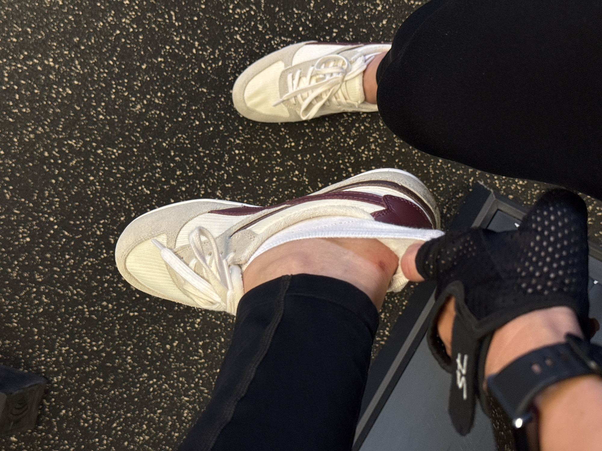 The image shows a close-up view of a person's legs and feet, wearing black leggings and athletic shoes. The person is sitting, with one foot placed on a black gym bench. The other foot is resting on the floor, which appears to be a textured, black rubber surface commonly found in gyms. A black weightlifting glove with mesh detailing is visible on the person's left hand, and a smartwatch is on their wrist. The background consists of the gym floor and a portion of gym equipment. There are no visible indications of the location being specifically in Sochi, Russia; it is a generic gym environment.