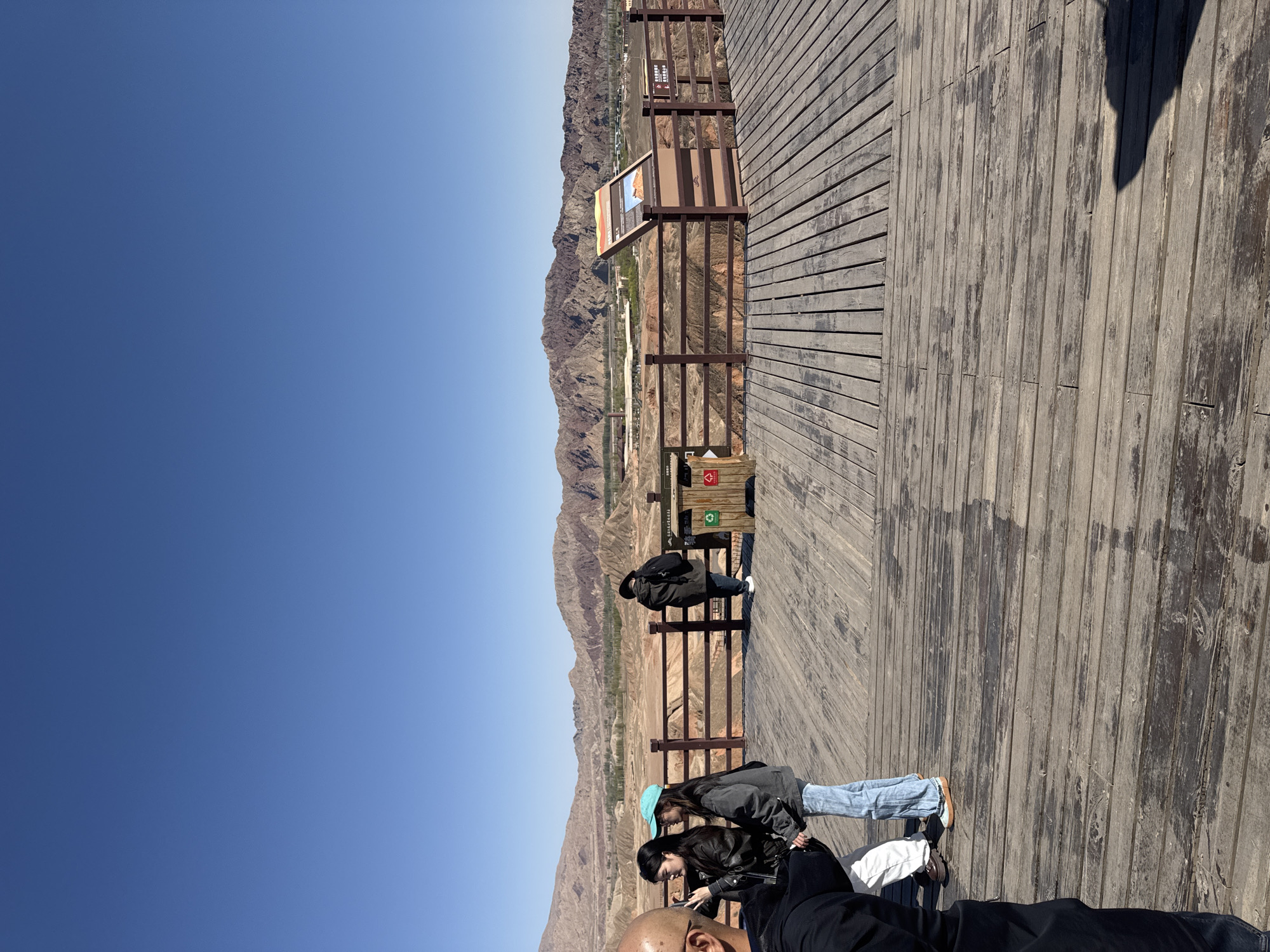 The image displays a scenic overlook in Zhangye, China, showcasing the vast, undulating Danxia landforms under a clear, bright blue sky. A robust wooden boardwalk occupies the foreground and mid-ground, serving as a viewing platform for visitors. A wooden railing delineates the edge of the platform, separating it from the expansive mountainous terrain below.
Four individuals are present on the boardwalk. In the immediate foreground, a person's bald head and dark-clothed shoulders are partially visible. Further back, two women stand close together; one wears a black jacket, blue jeans, and a light blue baseball cap, while the other is in a black jacket and black pants. They appear to be focused downwards, possibly at a device or map. In the background, a man leans casually against the railing, observing the panoramic view.
The landscape beyond the railing features the characteristic multi-colored strata of the Zhangye Danxia Geopark, with distinct layers of reds, browns, oranges, and grays forming the rolling hills and valleys. Hints of sparse vegetation are visible in some lower sections of the terrain.
The scene is set on a clear and sunny day, likely midday or early afternoon, as evidenced by the sharp shadows cast by the railing and people on the boardwalk. Several informational signs are mounted on wooden posts along the railing. While most text is illegible due to distance, one sign near the back clearly shows symbols for recycling and general waste. Another sign closer to the women displays a landscape image and some accompanying text.