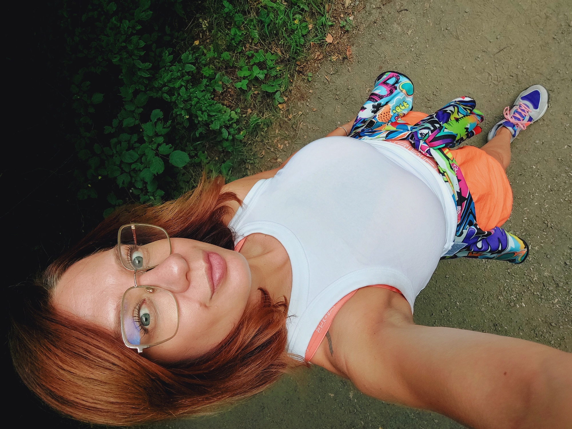 A woman with auburn hair and glasses stands on a dirt path, with greenery in the background. She is wearing a white tank top, orange shorts, and colorful athletic shoes. A vibrant, graffiti-style patterned garment is tied around her waist. The image appears to be taken from a high angle, likely a selfie. The location is identified as Bistritsa, Bulgaria.