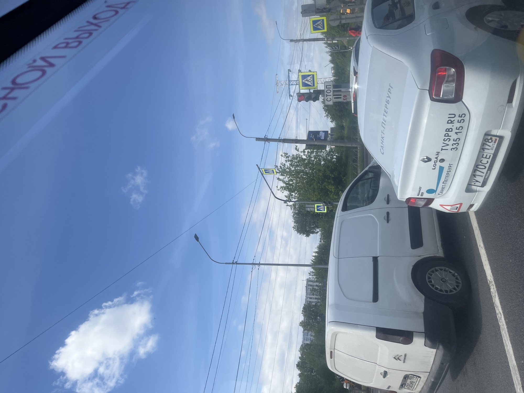 This vertical-format image, rotated 90 degrees counter-clockwise, depicts a bright daytime street scene in Saint Petersburg, Russia, from inside a vehicle. The sky, a clear blue with sparse white clouds, dominates the upper portion. Below, two white vehicles are visible on a paved road, apparently stopped or parked in traffic.
In the foreground, a reflection of red Russian text, "АВАРИЙНЫЙ ВЫХОД" (Emergency Exit), is visible on what appears to be a car's windshield. The vehicle in front is a white Renault Logan sedan, seen from the rear. Its rear window and trunk are covered with blue and black text. Key visible texts include "САНКТ-ПЕТЕРБУРГ" (Saint Petersburg) repeated multiple times, "TVSPB.RU", a phone number "335 15 55", and the car's model "LOGAN" and brand "RENAULT". The license plate reads "У 170CE 178 RUS", with "178" being the region code for Saint Petersburg.
Partially visible behind and to the left of the sedan is a white Citroën panel van, also seen from the rear. Its side door and a portion of the rear are visible, along with its brand name "CITROËN". A partially obscured license plate on the van reads "585MB 198", with "198" indicating a Moscow Oblast region code.
The street environment features numerous utility poles, overhead wires, and streetlights. Several traffic signs are visible, including multiple blue square signs with a white triangle and a walking person symbol, indicating pedestrian crossings. A traffic light shows a red signal, accompanied by a small vertical sign that reads "СТОП" (Stop). Green trees line the background, with distant buildings peeking through. The bright sunlight and blue sky suggest fair weather.