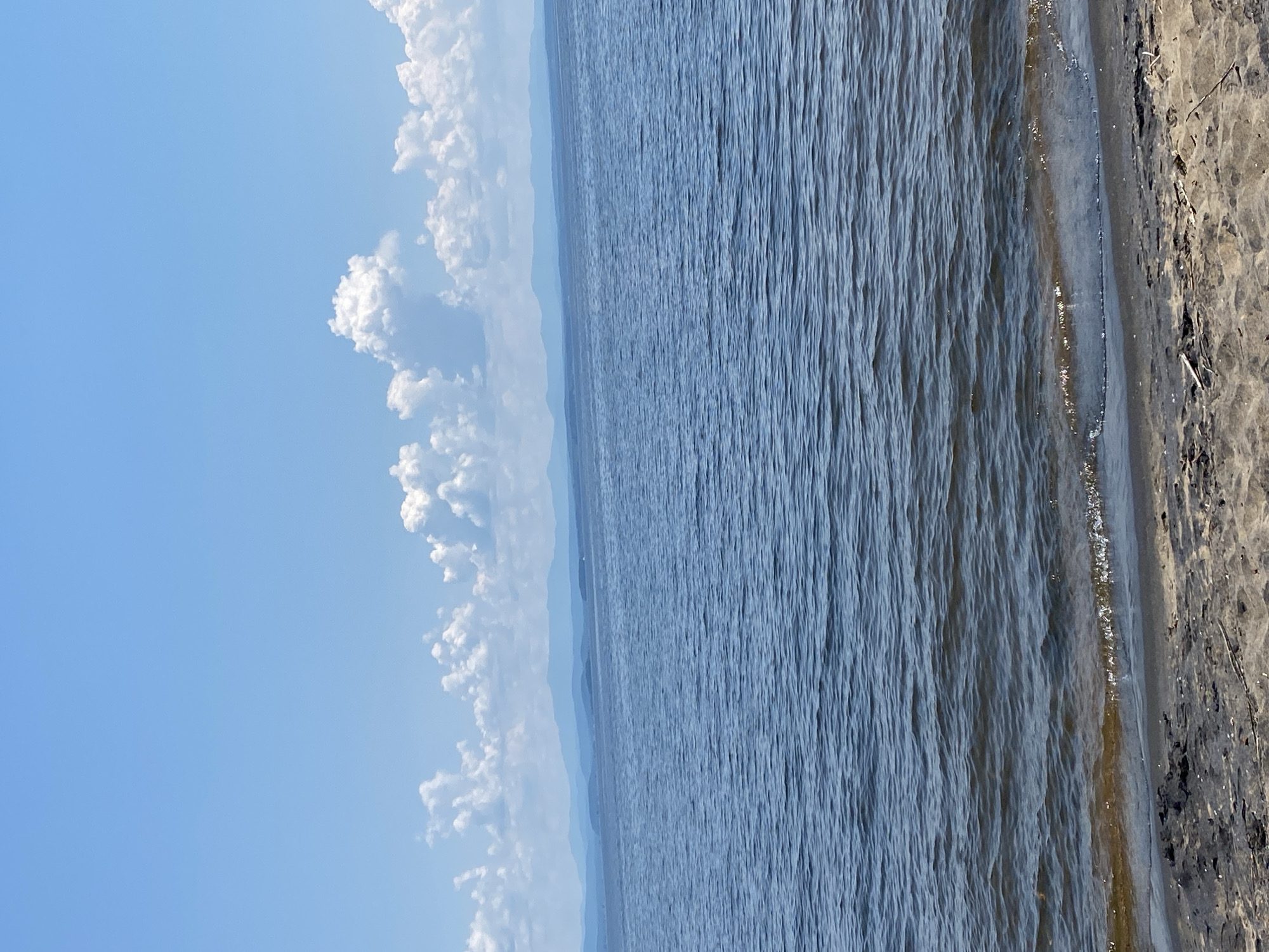 The image shows a tranquil scene of a body of water, likely a lake or ocean, bordering a sandy shore. The water is a deep blue, with gentle ripples creating a textured surface. In the distance, a hazy silhouette of land can be seen, suggesting a vast expanse of water. The sky above is a clear, bright blue, dotted with fluffy white clouds that form a striking contrast against the azure backdrop. The clouds are clustered along the horizon, adding depth and visual interest to the sky. The shoreline on the right side of the image is composed of light-colored sand, with some scattered debris, hinting at a natural, undeveloped setting. The overall impression is one of peace and serenity, with the natural elements of sky, water, and sand harmoniously coexisting. The lighting suggests it is daytime, likely mid-morning or late afternoon, given the soft quality of the light. The location is identified as the Town of Peru, United States of America. There is no text visible in the image.