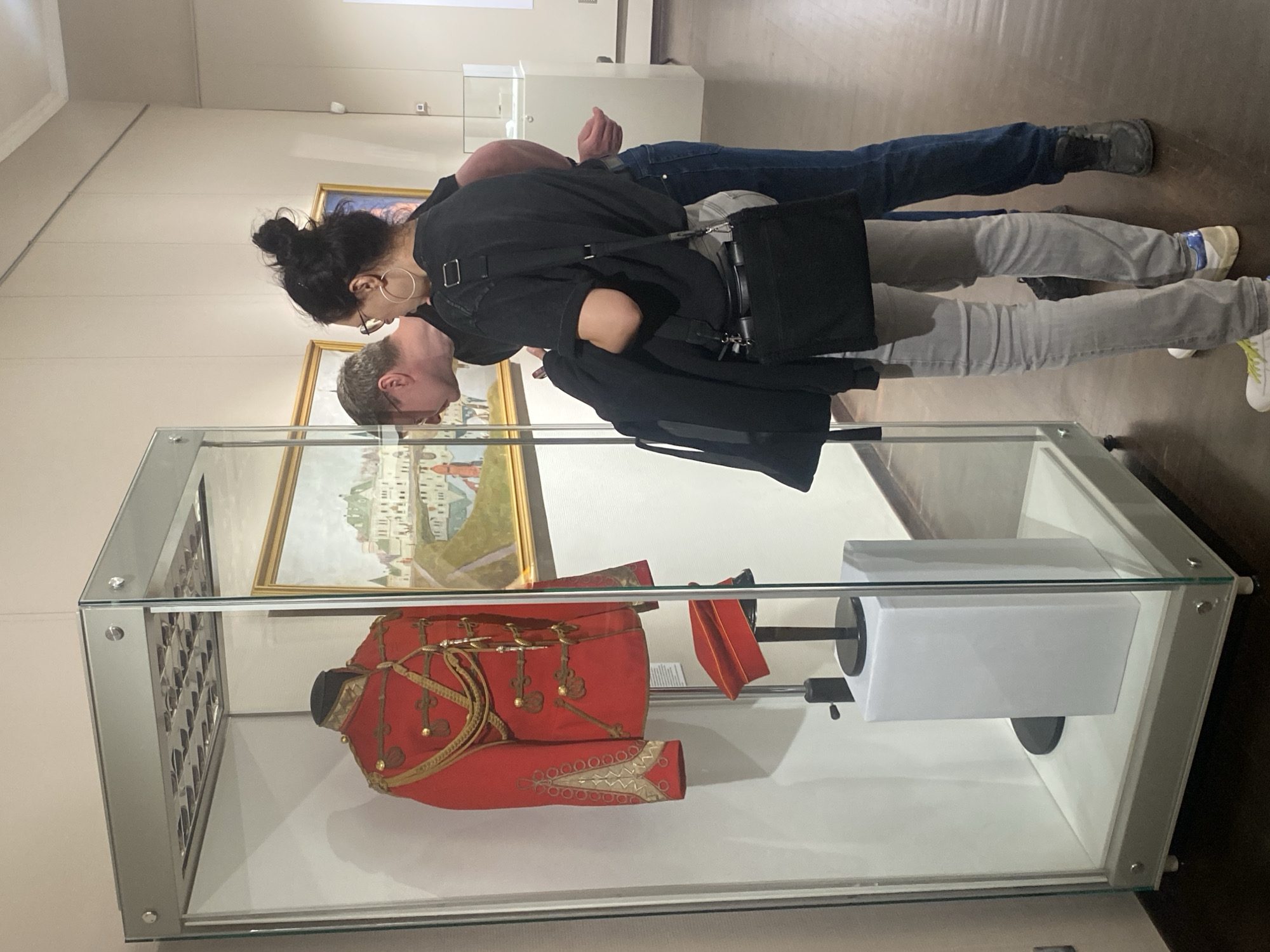 In Yekaterinburg, Russia, within a museum hall, a man and a woman are observing a display case. The display case contains a red military uniform adorned with gold trim and epaulets, along with a red cap positioned on a stand. The man, wearing glasses and a dark shirt, leans in to get a closer look. The woman, with her hair in a bun and wearing large hoop earrings, a dark t-shirt, and light grey jeans, stands beside him. A black bag is slung across her body. Behind them, another person is partially visible, wearing blue jeans. The museum walls are a neutral color, and paintings are visible on the wall in the background. The lighting in the hall appears to be from overhead fixtures.