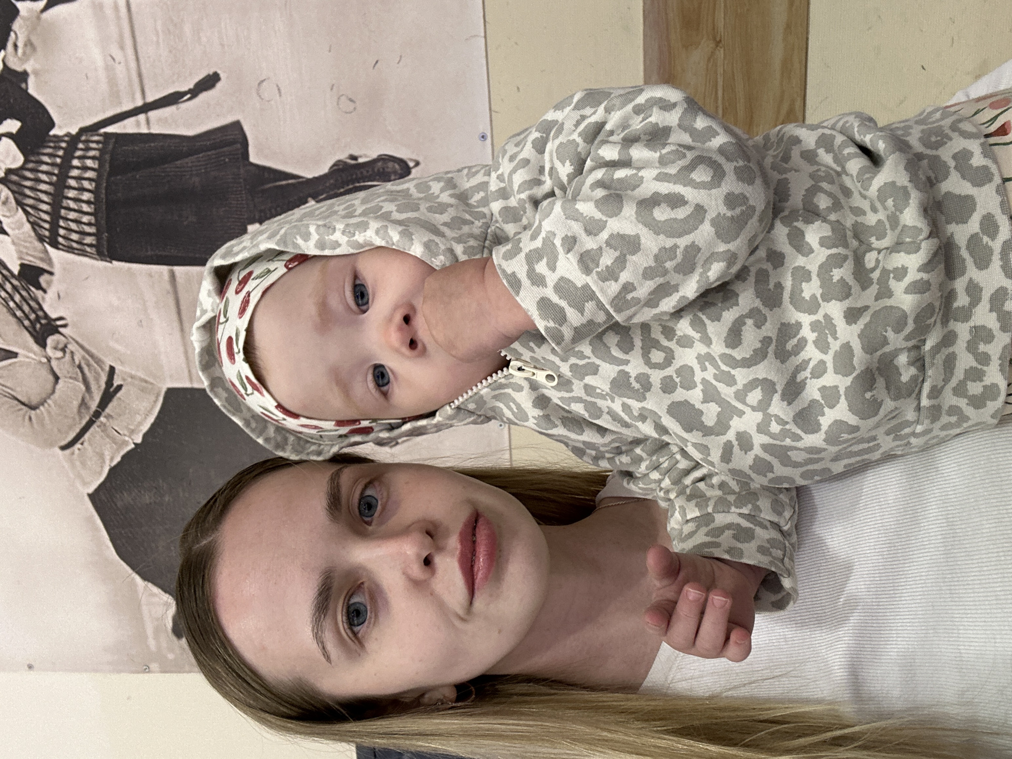 A medium shot depicts a blonde woman lying on a white surface, possibly a bed, with a baby lying on top of her. The baby wears a grey leopard-print hooded sweatshirt and a headband with a cherry pattern. The baby's finger is in their mouth. A framed photograph of a woman in a black-and-white period dress is visible in the background. The image was likely taken indoors in Chaikovsky, Russia.