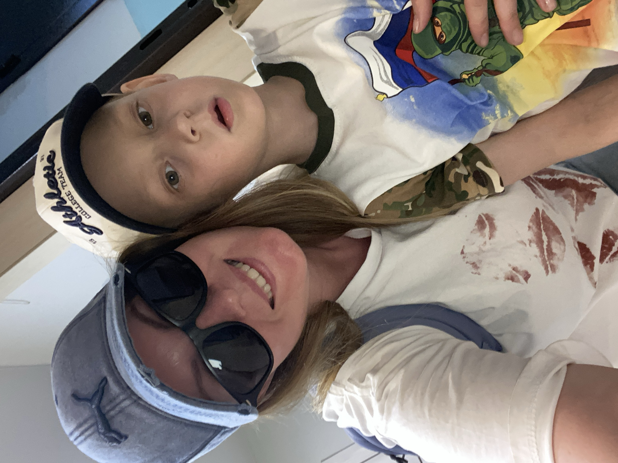 A woman and a boy are in the frame. The boy lies on the woman. The woman is wearing a blue hat and sunglasses. She's wearing a white t-shirt with red lipstick kiss marks. The boy is wearing a hat and a t-shirt with a cartoon ninja on it. The location cannot be determined from the image.